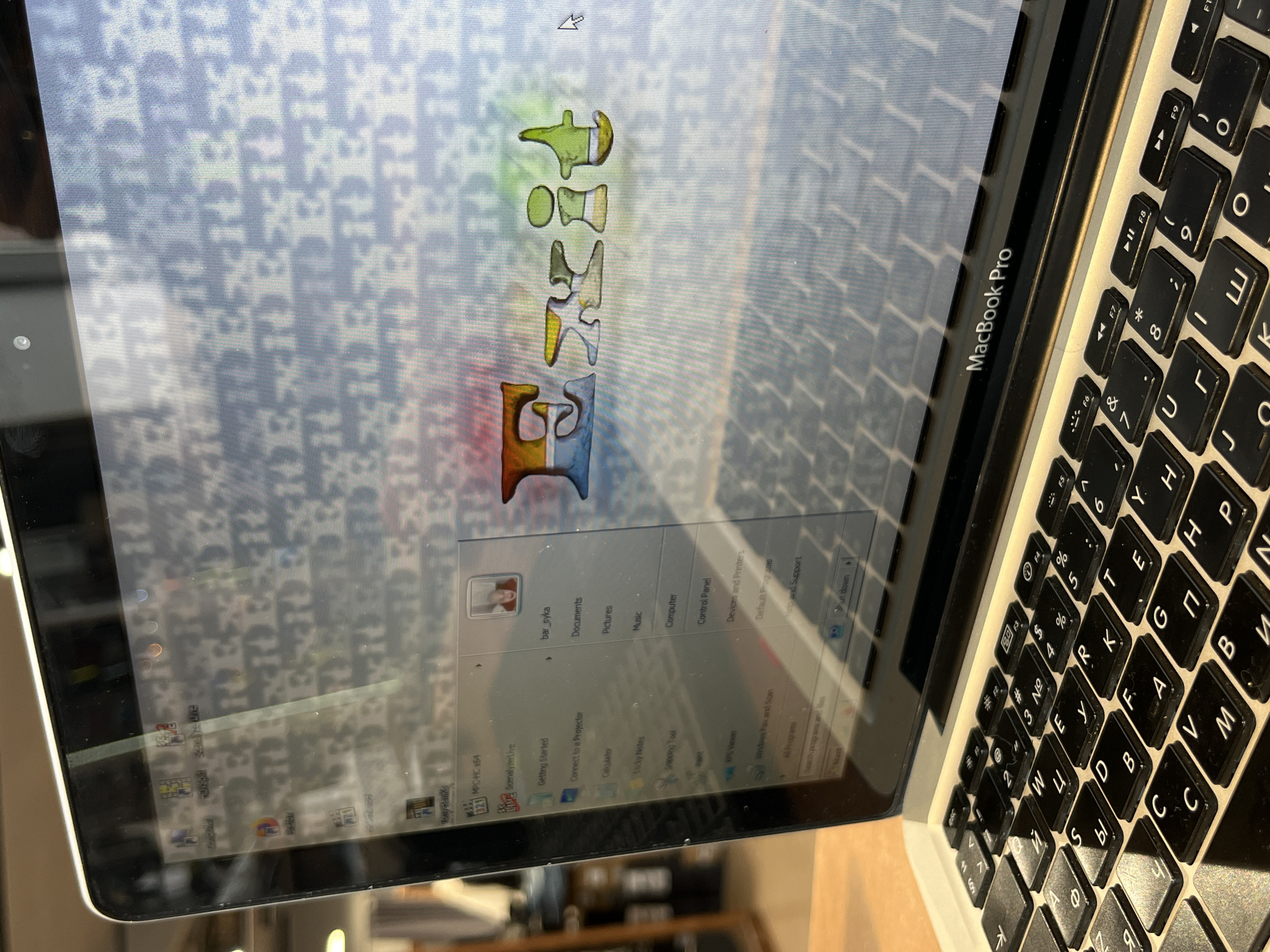 The image shows a MacBook Pro laptop displaying the Windows XP operating system. The desktop background features a pattern resembling the Dior logo. An open window titled "bar_syka" suggests a user profile or file. The laptop is likely located in Krasnoyarsk, Russia, given the Cyrillic characters on the keyboard and the presence of the "bar_syka" user profile.