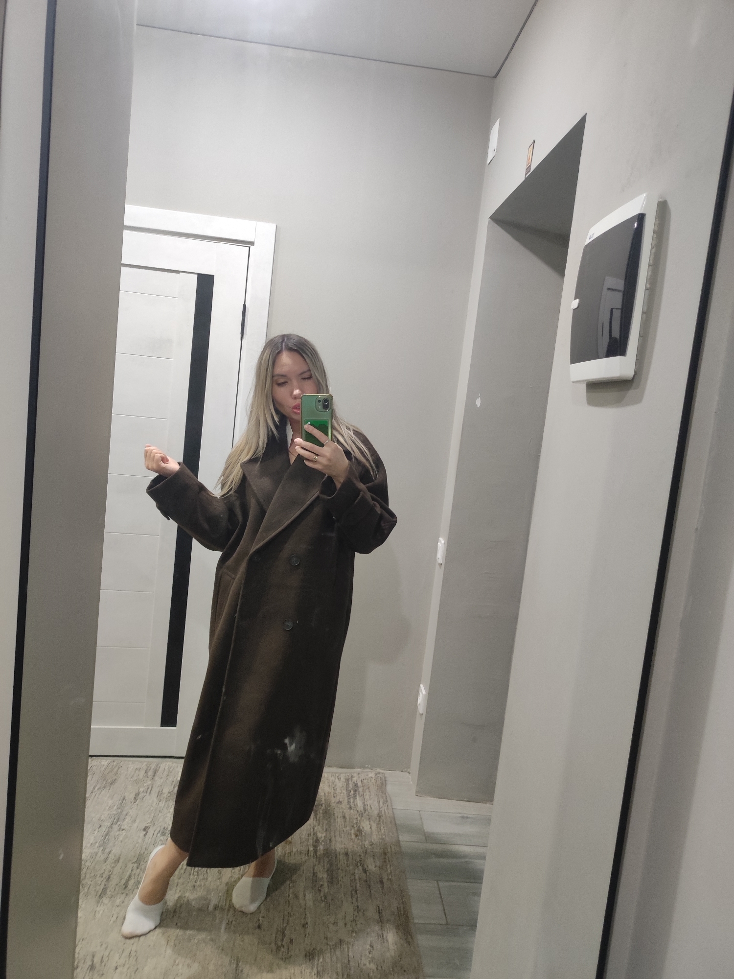 A woman stands in front of a mirror taking a picture of herself. She is wearing a long, brown, double-breasted coat and white shoes. Her blonde hair is long and straight. She holds a green smartphone. The room appears to be an entryway with gray walls. There is a white door behind her, and a doorway with a wall-mounted device to the right. The floor has tile. The location is not specified as Votkinsk, Russia.