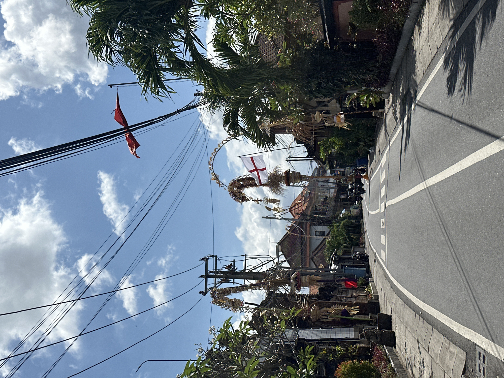 The image depicts a street scene in North Kuta, Indonesia, under a bright blue sky with scattered white clouds. The road is asphalt with white lane markings, curving to the right in the foreground. On the left side of the road, a sidewalk with a decorative border is visible, lined with greenery. Traditional Balinese decorations, made of dried leaves and woven materials, are prominently displayed, attached to utility poles and arching over the street. One such decoration on the left features a tall, curved structure resembling a serpent's head, adorned with smaller woven elements. Further down the street on the right, a white flag with a red cross is flying from a similar decorative arch. Palm trees and lush foliage flank the buildings lining the street, which appear to be a mix of residential and commercial structures with tiled roofs. Utility wires are strung across the sky, crossing the frame at various angles. Several motorcycles are parked along the roadside and a few are in motion on the street, indicating ongoing activity. A red flag flutters in the wind from a pole near the utility wires.