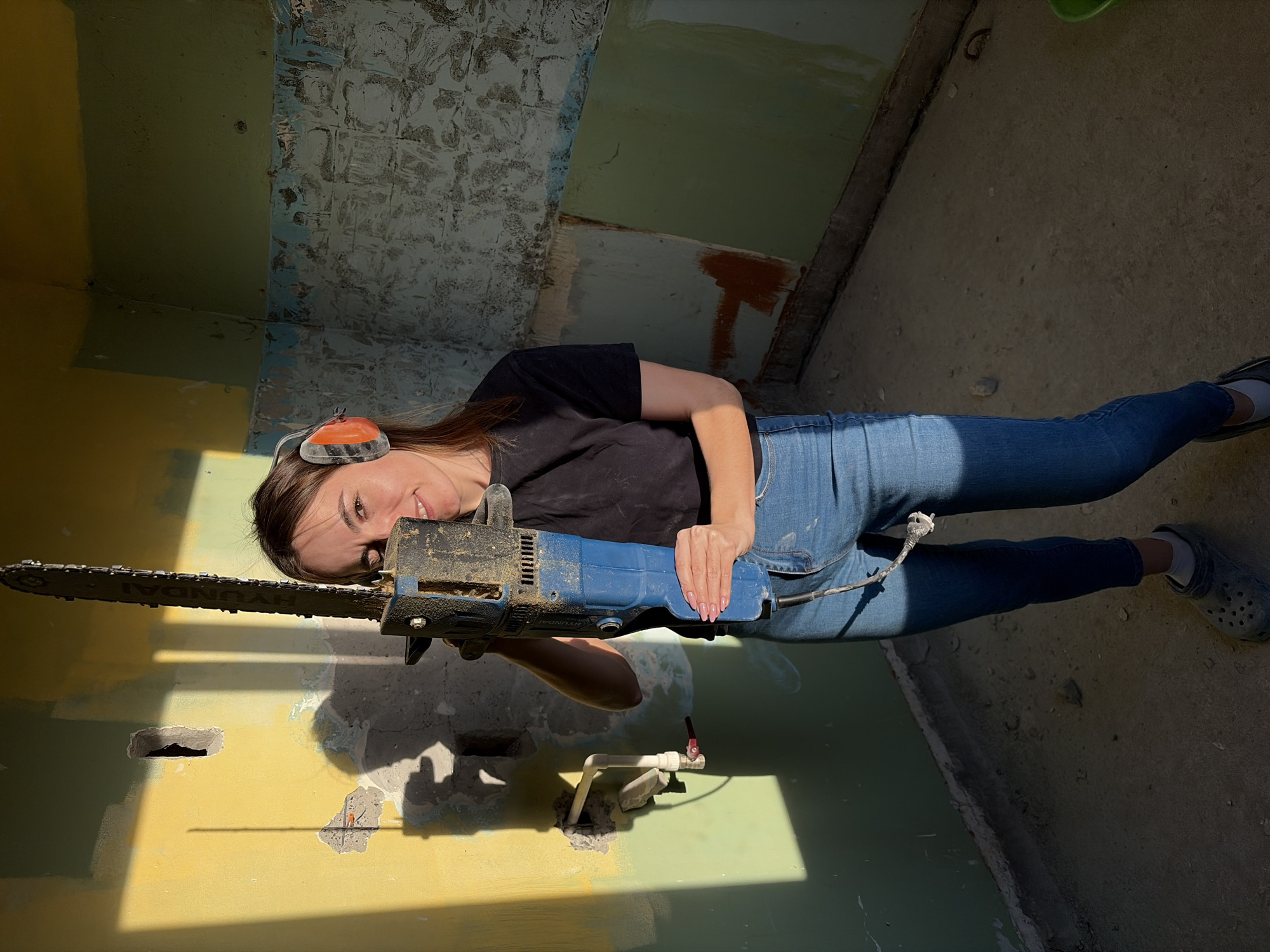 A woman is indoors, posing with a demolition chainsaw in an interior space that appears to be undergoing renovation. She is wearing blue jeans, a black t-shirt, safety earmuffs, and Crocs. She is holding the chainsaw with both hands, directing the blade towards the viewer. The wall behind her is painted in sections of yellow, green, and blue, with areas of damage and exposed surfaces. A pipe protrudes from the wall. The floor appears to be concrete. The photo was not taken in Izhevsk, Russia.