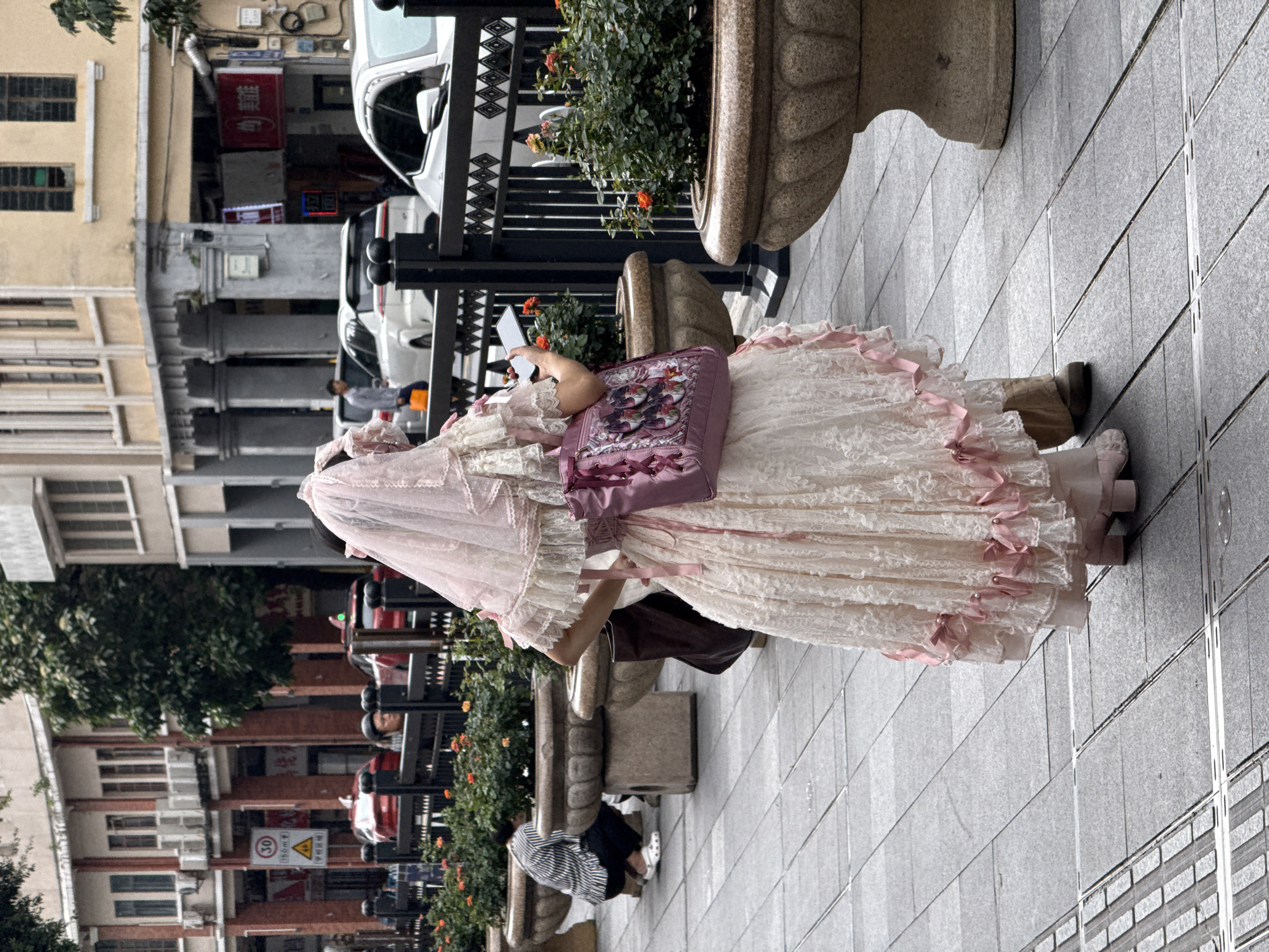 A person in an ornate, frilly pink and white dress stands on a grey tiled sidewalk next to a stone planter with flowers. The person is holding a smartphone. A pink bag is slung over their shoulder. The location is Yuexiu District, China. The street has buildings on both sides. One side shows vehicles parked along the street and a building with a storefront. The other side shows older buildings and trees. There are also pedestrian benches with black wrought iron features.