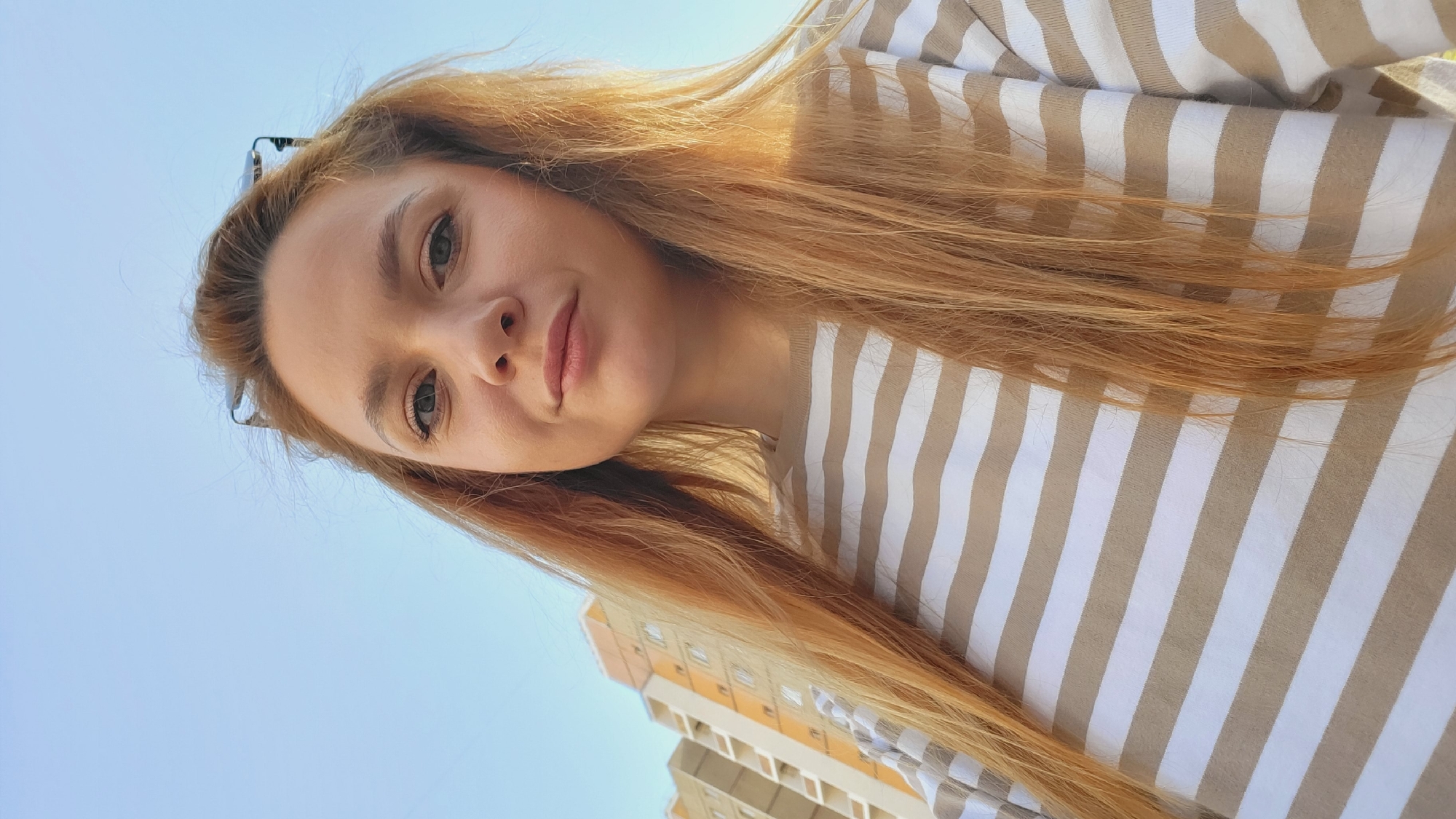 A young woman with long, strawberry blonde hair poses for a selfie in Chaikovsky, Russia. She wears a beige and white striped shirt. Behind her is a clear blue sky and a multi-story apartment building with beige and orange panels. She has glasses perched on top of her head. The perspective is from below, looking up at the woman.