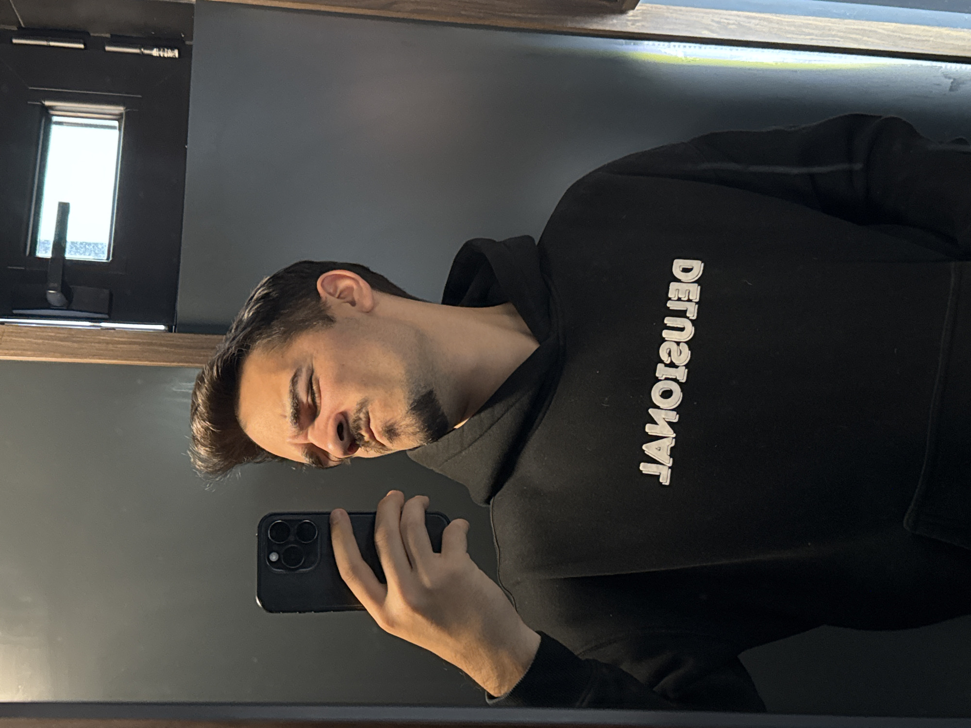 A man is shown in a vertically oriented medium shot, presumably a reflection in a mirror. He is holding a black smartphone in his left hand, seemingly taking a photograph of himself. The man has fair skin, dark hair styled with a slight wave or comb-over, and a neatly trimmed mustache and beard. He's wearing a black hooded sweatshirt with the word "DEJAOITZEL" printed in white letters backwards across the front. The background is a neutral gray wall, with a window visible in the upper left corner. There is no evidence to determine if the image was taken in Sofia, Bulgaria.