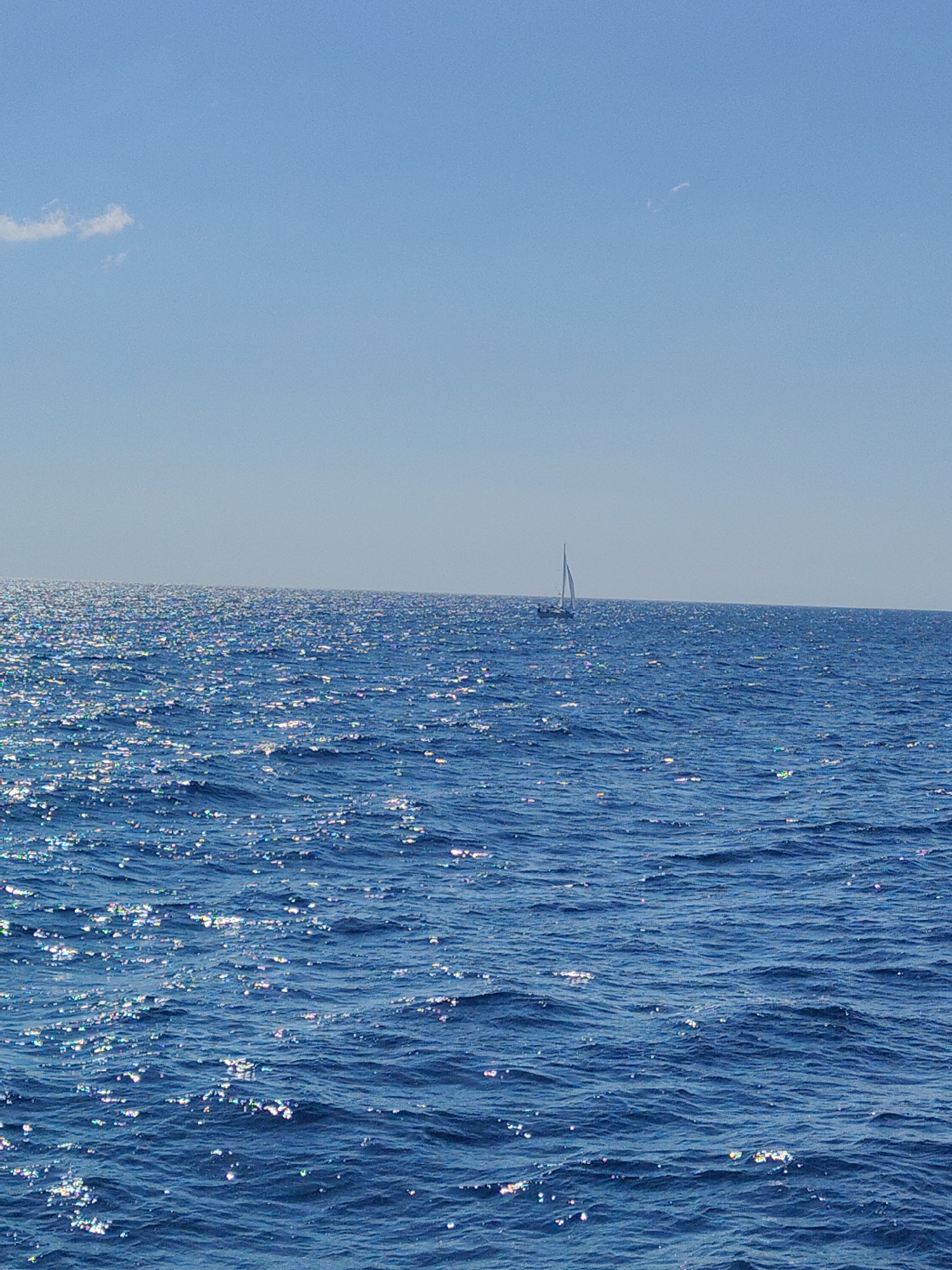 A sailboat with a white sail is visible on the horizon, set against a clear blue sky and a vast expanse of shimmering blue ocean. The sun reflects brightly off the water, creating a sparkling effect. A small, wispy cloud is present in the upper left portion of the sky. The scene appears to be a tranquil day at sea, possibly near the coast of Sipias Municipal Unit, Greece, known for its scenic maritime landscapes.