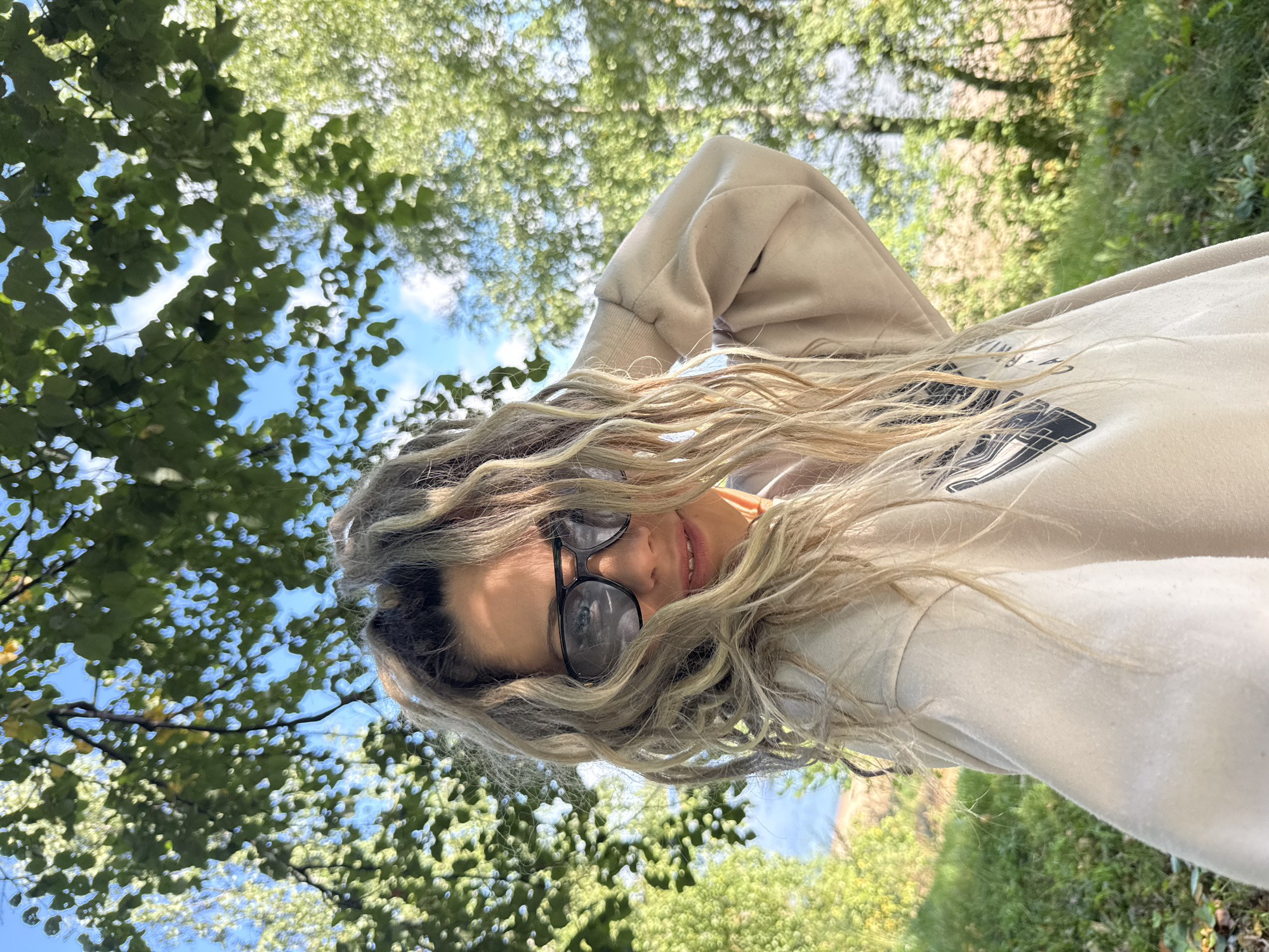 A young woman with long, blonde, wavy hair and sunglasses is taking a selfie outdoors. She is wearing a light-colored sweatshirt. The background is filled with green trees and a blue sky with some clouds, suggesting a sunny day. The ground is covered in green grass. The scene appears to be a park or natural area within or near the city of Rusanovo, Russia.