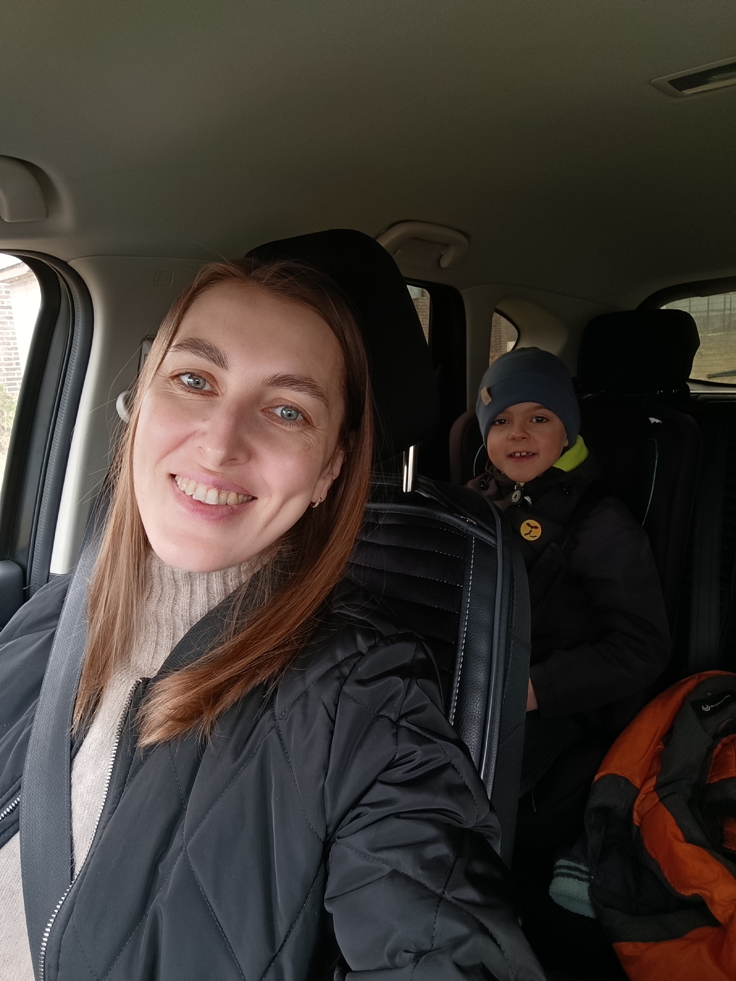 A heartwarming selfie captures a woman and a young boy inside a car, likely in Chaikovsky, Russia, on a daytime outing. The woman, positioned in the front passenger seat, smiles broadly at the camera, her bright blue eyes and long brown hair framing her face. She wears a light-colored, ribbed turtleneck sweater underneath a black quilted bomber jacket, with a grey seatbelt securely fastened across her chest. A subtle earring is visible in her right ear.
In the background, a young boy is seated in a car seat, positioned behind the front passenger. He also smiles, revealing some front teeth, and is dressed warmly in a dark blue or grey beanie and a dark, possibly black, jacket with a visible lime green or yellow lining near the neck. A circular yellow pin with a black bat-like emblem is affixed to his jacket. Both subjects appear happy and content.
The car's interior is visible, featuring dark seats and a light grey headliner. Through the windows, blurred views of what appear to be residential buildings or structures with light-colored facades can be seen, suggesting an urban or suburban setting. An orange and grey jacket or bag is visible in the footwell in the lower right corner, next to the child. The overall scene suggests a casual journey or outing.