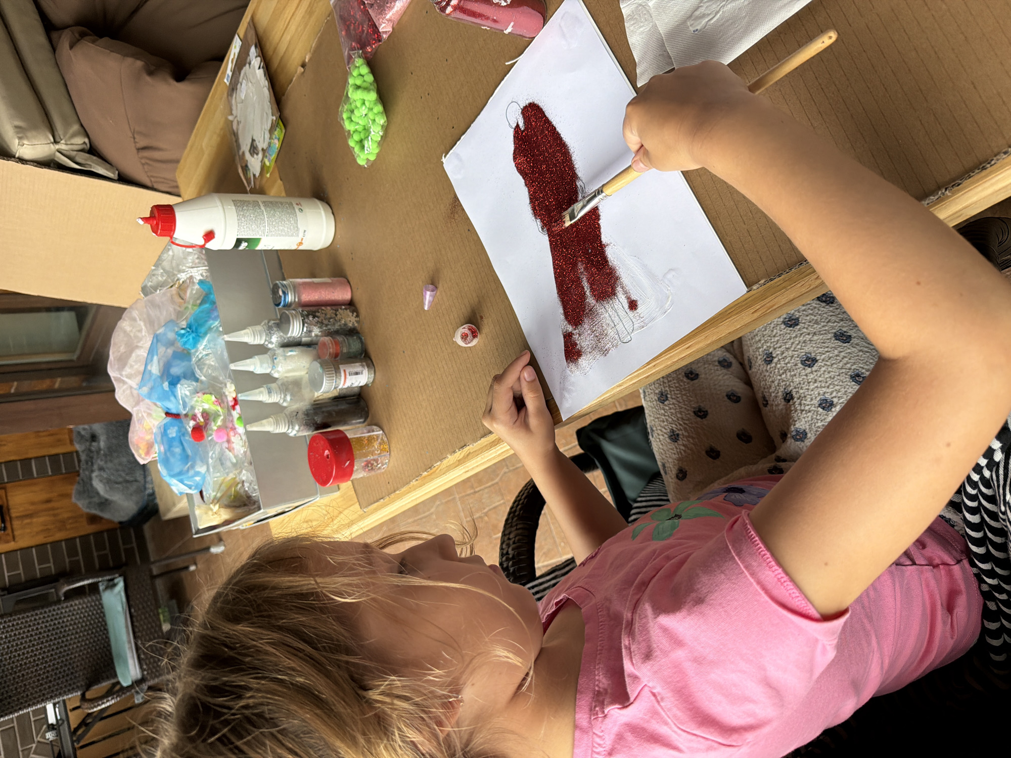 A young girl with blonde hair, wearing a pink t-shirt, sits at a wooden table outdoors and is applying red glitter to a drawing with a paintbrush. The table is covered with craft supplies, including bottles of glitter, glue, and pom-poms. In the background, there is a chair, a large cardboard box, and some cushions. The scene appears to be in a backyard or patio area.