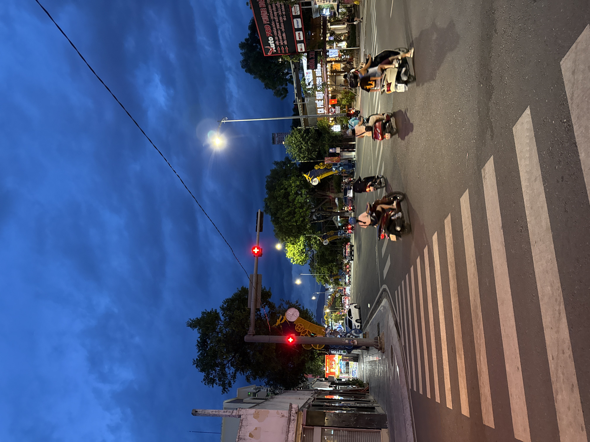The image depicts a street scene at dusk in Nha Trang, Vietnam, with a cloudy, dark blue sky overhead. The perspective is from a pedestrian crossing, looking towards an intersection. On the left side of the street, there is a sidewalk with buildings lining it, some with their shutters down. A traffic light with a red signal is visible.
The road is paved and features a pedestrian crossing with white stripes. Several people are riding motorbikes, navigating the intersection. The motorbikes are in motion, and their headlights and taillights are illuminated. Some individuals on the motorbikes appear to be passengers.
In the background, there are more buildings, trees, and streetlights. A tall lamppost with a bright light stands prominently in the middle ground. A large billboard advertising "Auto 789 PHUONG HOA" is visible on the right side of the road, along with other signs for businesses like "CA PHE HAT" and "RUOU XE". The overall atmosphere is that of a busy street at twilight, with the city lights beginning to illuminate the scene.