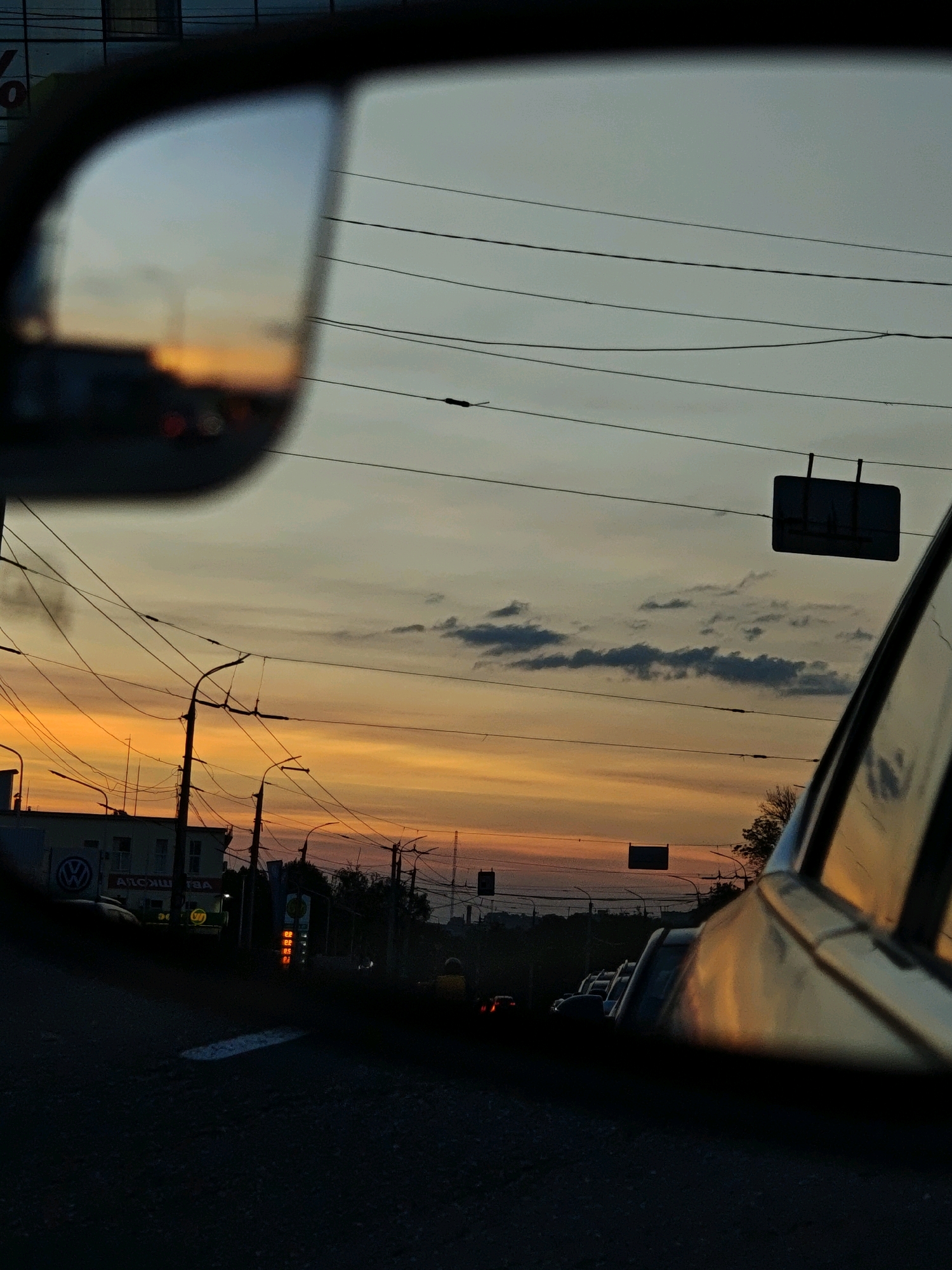 This image captures a sunset viewed from the perspective of someone in a vehicle on a street in Vinnytsia, Ukraine. The sky is a vibrant mix of orange and yellow hues, with scattered clouds. Overhead power lines crisscross the sky, typical of an urban environment. In the foreground, the side mirror of the vehicle reflects a portion of the scene, showing the colorful sky and some blurred traffic lights. On the left side of the image, buildings and streetlights are visible, suggesting a commercial or residential area. The road surface in the foreground is dark asphalt, indicating it is likely nighttime or dusk. The overall impression is a city scene bathed in the warm glow of a sunset.