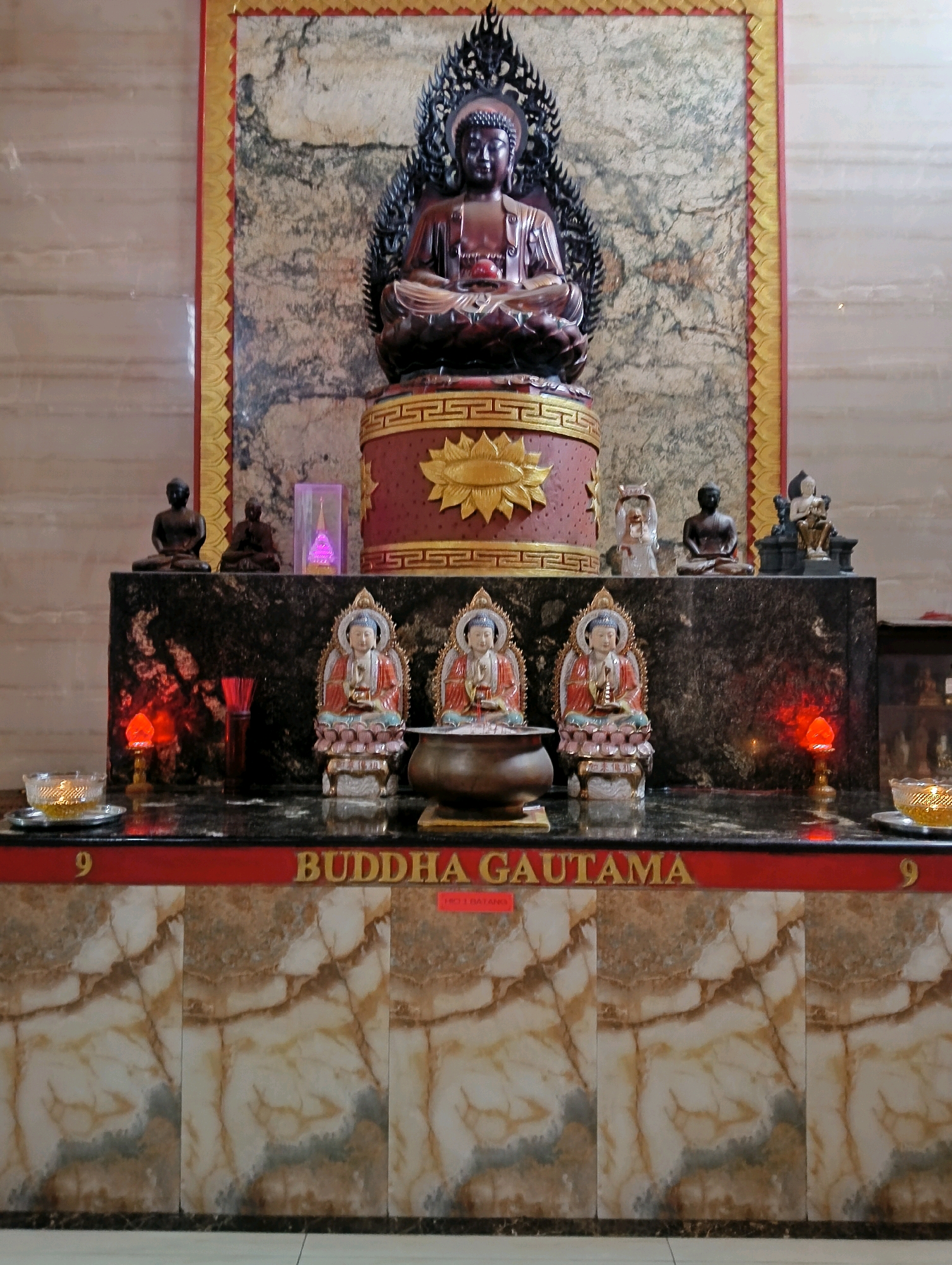 This is a photograph of a Buddhist altar, likely located within a temple or shrine. The main subject is a large, dark statue of Buddha seated in meditation on a tiered, ornate pedestal. The pedestal is red with gold accents, featuring a sun-like motif. To the left and right of the main Buddha statue, on a lower level, are smaller statues of meditating figures. In front of the main statue, on a dark marble surface, are three smaller, colorful Buddha statues, also seated in meditation. A bronze bowl sits in front of the central smaller Buddha. Red and orange lights are visible, illuminating the altar. The backdrop of the altar features a textured wall with a gold and red border. Below the altar, a red band displays gold lettering that reads "9 BUDDHA GAUTAMA". The lower portion of the image shows a wall with a marble-like pattern. The overall scene suggests a place of worship or religious significance. The location of this specific altar is not discernible from the image itself.
