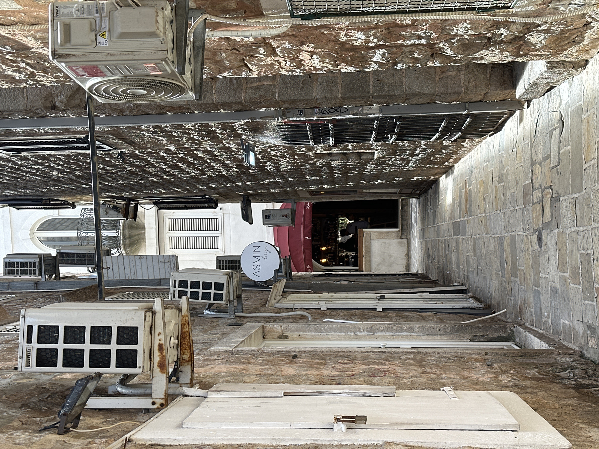 The image shows a narrow alleyway in Budva, Montenegro. Overhead are air conditioning units mounted on stone walls, some attached to the building façades and some suspended from the ceiling. The alley floor consists of stone slabs and ledges, interspersed with what appears to be access covers. A sign with "ASMIN design" is visible on one side. The alley leads to a dimly lit interior space. The photo is taken at an odd angle, with the top of the image seeming to show the ceiling of the alley. The overall impression is one of a somewhat rundown and utilitarian space.