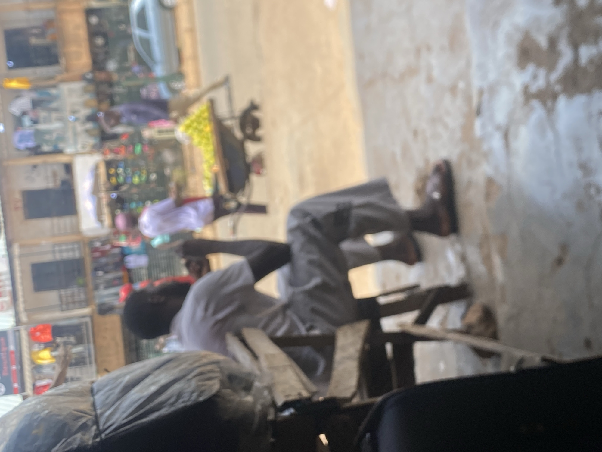 The image captures a scene in Bukuru, Nigeria. A man is sitting on a wooden chair outdoors. He is wearing light-colored pants and a light-colored shirt. To his left, there is a covered object. In the background, a street scene includes shops and a vehicle. A bicycle with fruit is positioned near him. The focus is somewhat blurred.