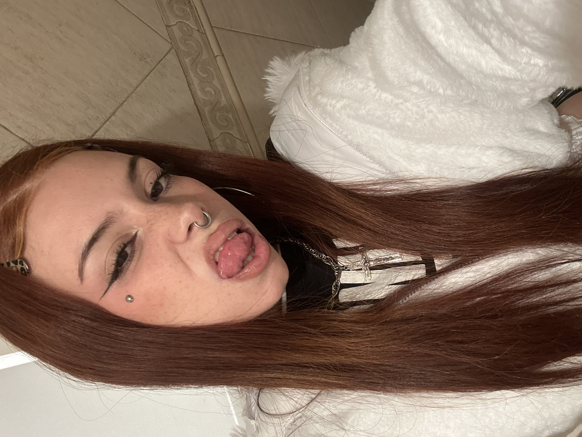 A woman is posing for the camera. She has reddish-brown hair and fair skin. She is wearing a nose ring and has a piercing under her left eye. She has a black eyeliner wing. She is sticking her tongue out. She is wearing a white, furry jacket and a black shirt. A silver cross necklace is visible. The background includes tile and a decorative border. The location appears to be indoors. The image provides no information about the city of Buenos Aires, Argentina.