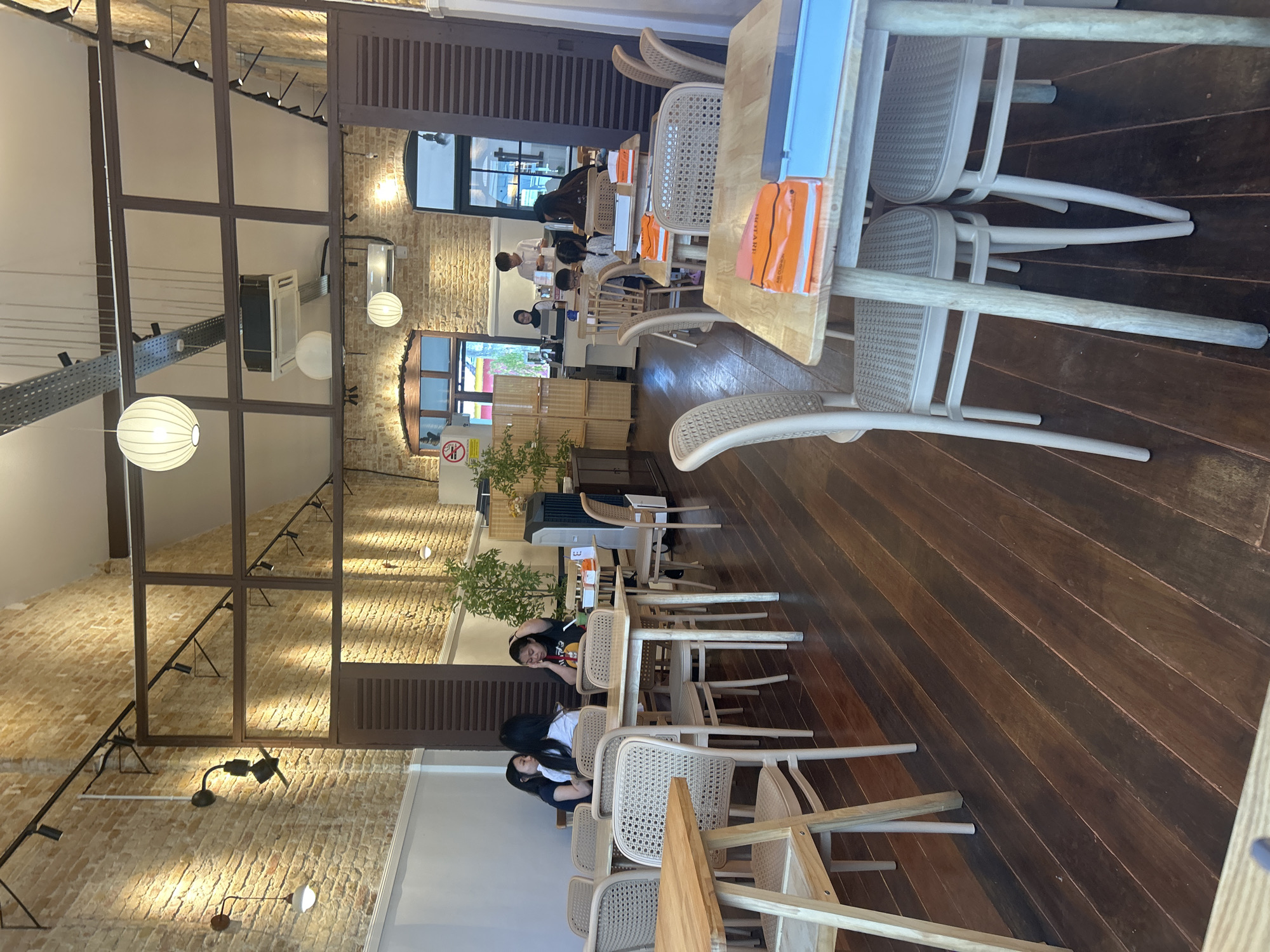 The image depicts the interior of a cafe, likely located in Ipoh, Malaysia, with a rustic and airy ambiance. The walls are exposed brick, and the ceiling features exposed metal ducting and track lighting. Several tables and chairs are arranged throughout the space, most made of light-colored wood and cane.
In the foreground, a table is set with orange and pink placemats and a blue box. To the left, another wooden table and chairs are visible. Further into the cafe, several patrons are seated at tables. On the left, three women are seated at a table, two facing away from the camera. In the middle and right sections, various other tables are occupied by individuals and groups, including a server standing near a counter in the background. Plants are placed in decorative pots, adding a touch of greenery to the space. The floor is made of dark, polished wooden planks. The overall scene suggests a casual dining or coffee shop environment.