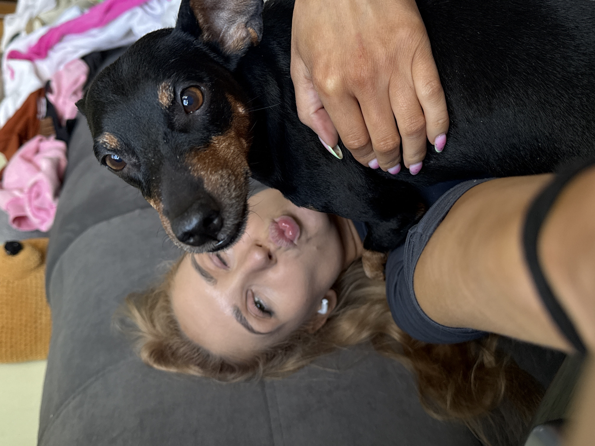 A woman lies on a gray, quilted surface, possibly a bed or sofa. She has blonde hair and is wearing a small white wireless earbud in her left ear. She is making a kissing face. A black and brown dachshund lies on top of her, its body mostly covering her torso. A woman's hand is visible on the dog's back. In the background, there is a pile of clothes including a pink garment. To the left, a stuffed bear is partially visible. No evidence indicates the location is Chaikovsky, Russia.