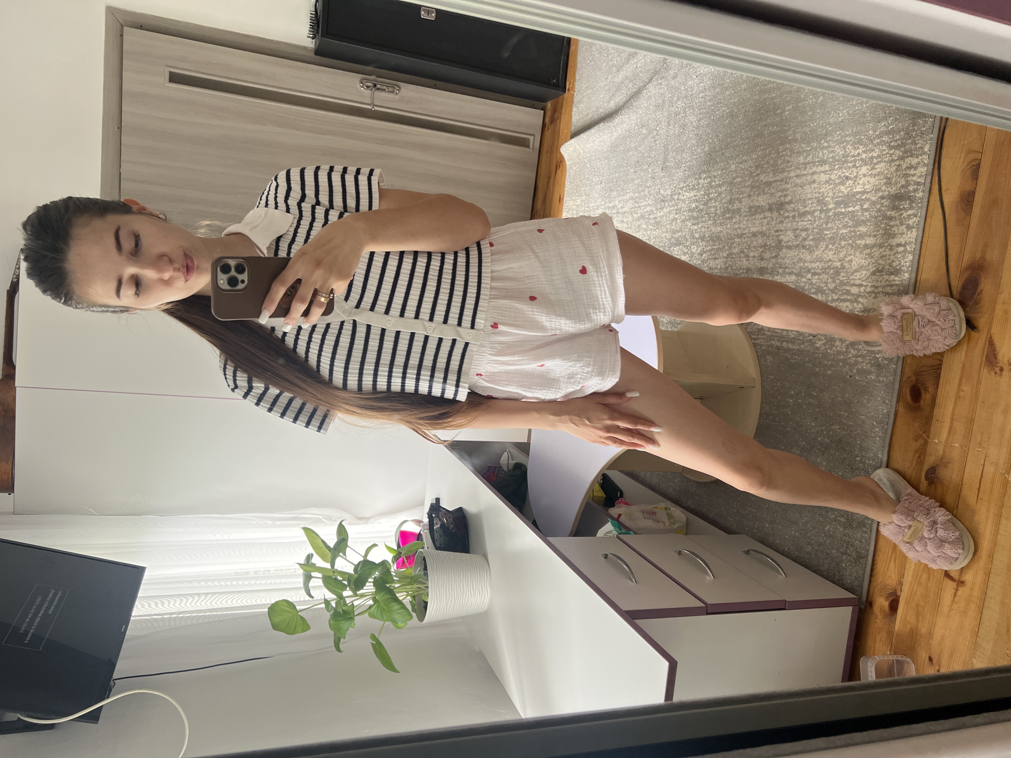 A young woman stands in a room, taking a selfie in a mirror. She has long, dark brown hair pulled back into a ponytail. She is wearing a striped short-sleeved shirt with a white collar and white shorts with a strawberry pattern. She is also wearing pink fuzzy slippers. To her left, there is a white desk with a potted plant on it, and a television mounted on the wall above. To her right, there is a closet door and a black speaker. The floor is made of wood planks, and there is a rug in front of the desk.