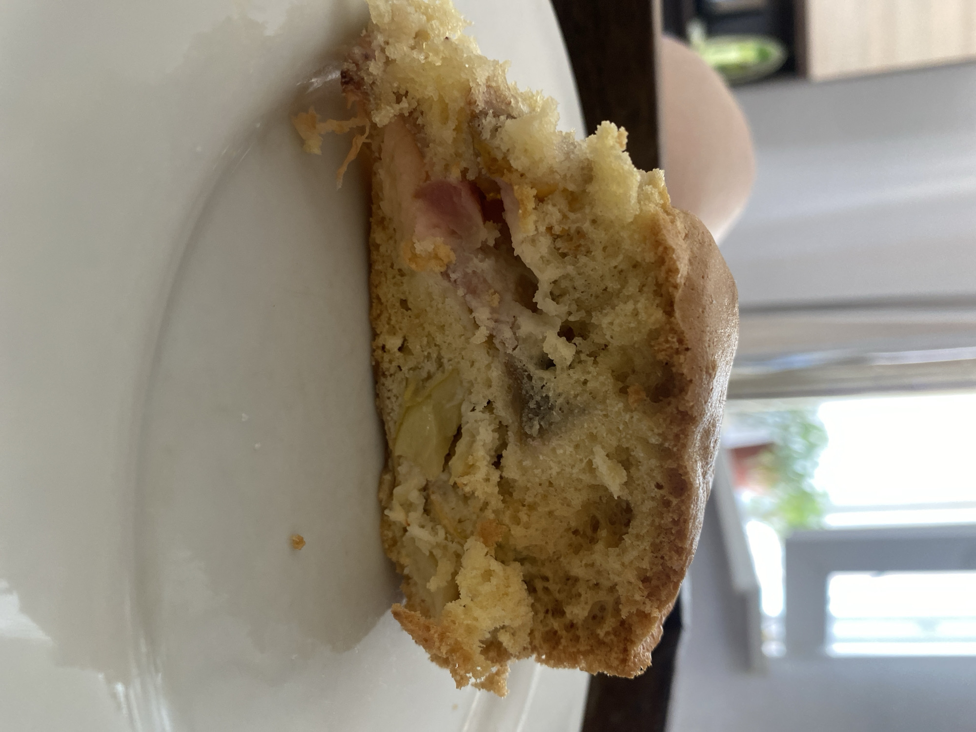 A slice of apple pie sits on a white plate, partially obscuring a person's knee and thigh in the background. The pie has a golden-brown crust and is filled with visible pieces of apple. The scene appears to be indoors, possibly in a kitchen or dining area, with natural light coming from a window in the background. The environment is casual and domestic, with no specific identifiable landmarks of Chornomorsk, Ukraine.
