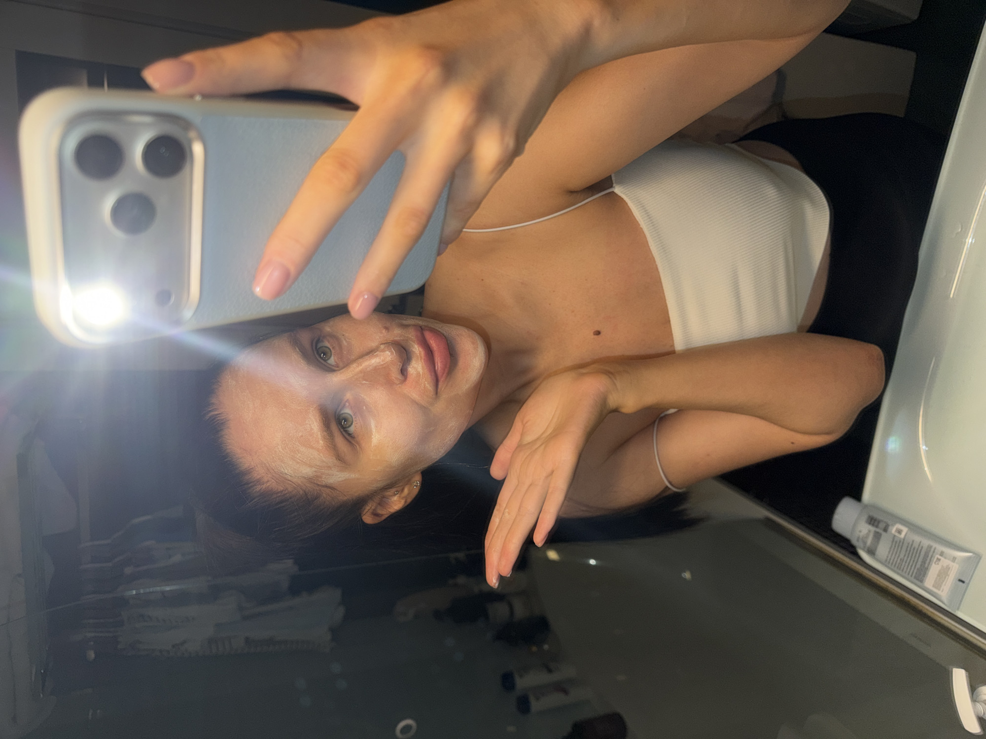 An indoor selfie shows a young woman in what appears to be a bathroom or vanity area, captured from a rotated perspective. She is holding a light-colored smartphone, identifiable as an iPhone with a triple-lens camera system and a prominent flash that illuminates the scene. Her nails are manicured with a light pink polish.
The woman is engaged in a self-care activity, as evidenced by a light-colored face mask applied to her skin, covering parts of her face. She wears a white ribbed, strappy top. Her expression is neutral as she looks directly towards the phone's camera. A small mole is visible on her upper chest.
The background includes a dark, highly reflective surface below her, showing indistinct reflections of objects and her face. To the right, a white sink or countertop is visible, on which a tube of cosmetic product rests. The text on the tube is largely unreadable, though generic labels like "ML" are discernible. The strong artificial light from the phone's flash suggests the photo was taken in the evening or at night. The location context is Khimki, Russia.