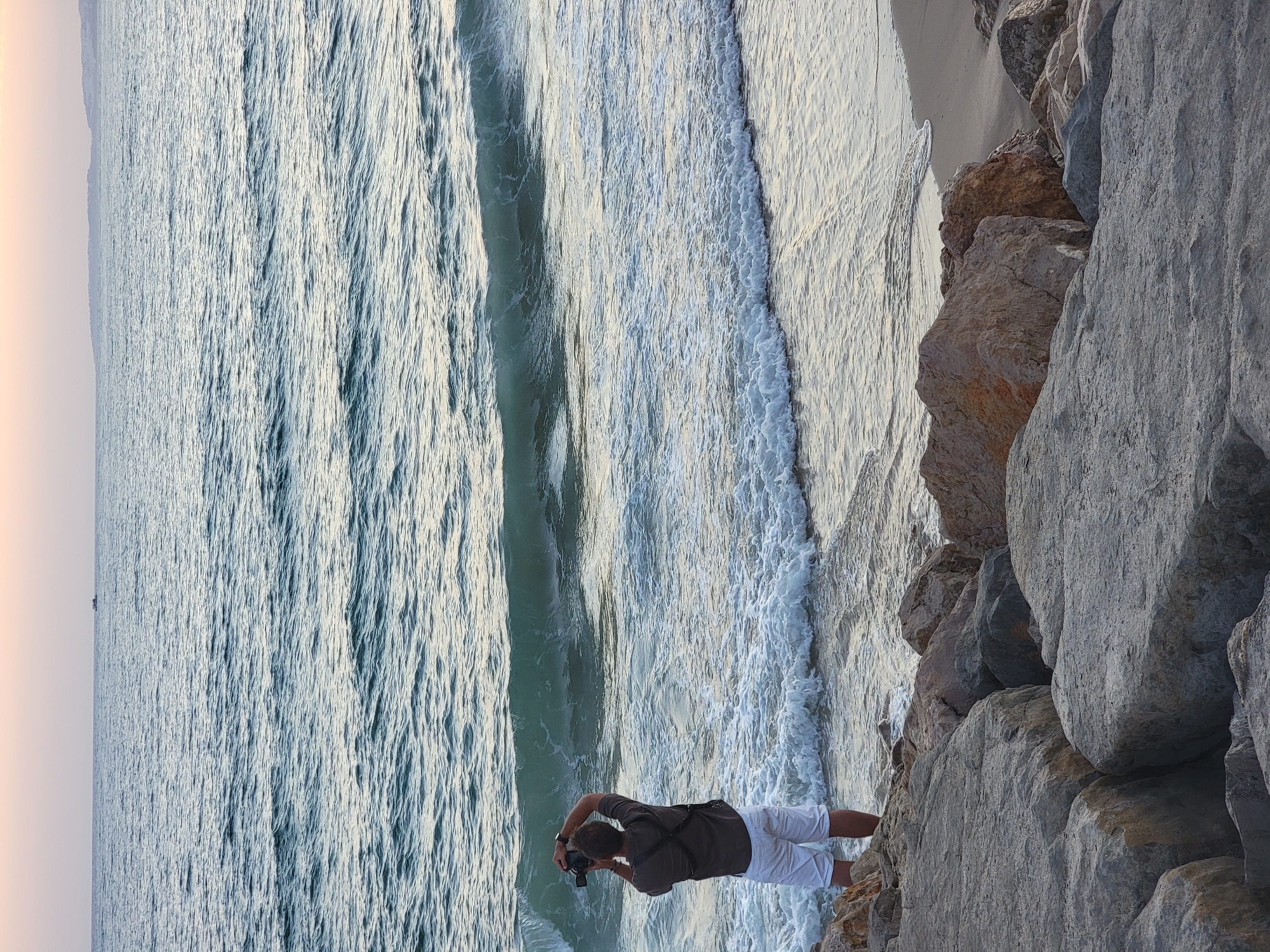 A lone man stands on a rocky shore, facing the ocean, in Quarteira, Portugal. He is wearing a dark shirt and white shorts and is holding a camera up to his eye, apparently taking a photograph of the waves. The ocean is calm with small waves lapping at the shore. The sky is a soft pink and orange, indicating either sunrise or sunset. In the distance, a small boat can be seen on the horizon. The foreground of the image is dominated by large, grey rocks, typical of the coastline in Quarteira. The scene evokes a sense of tranquility and contemplation.