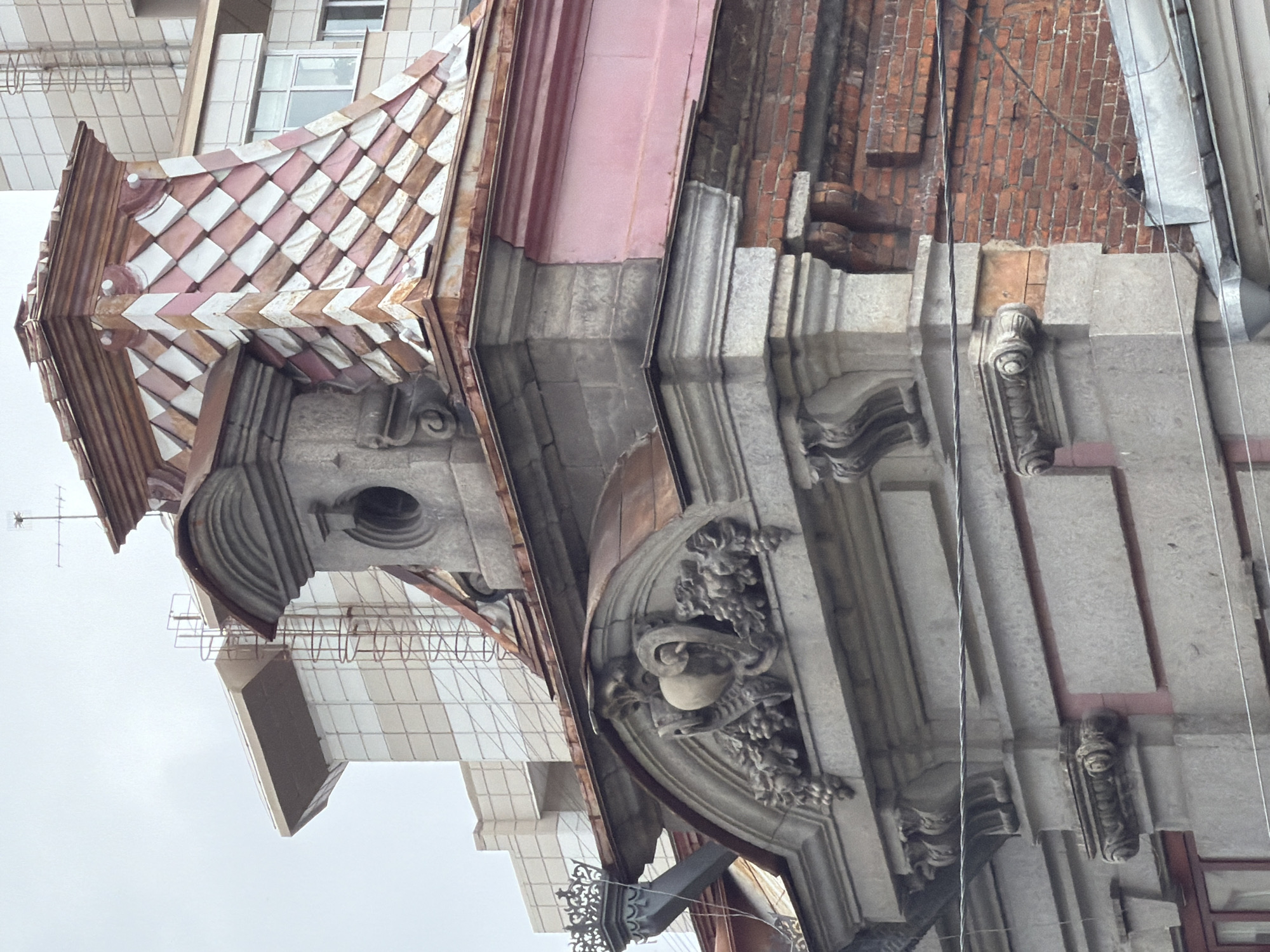 The image captures a detailed view of the upper facade of an old, ornate building in Yekaterinburg, Russia. The architectural style suggests a historic building, likely from the late 19th or early 20th century, characterized by decorative stonework and a distinctive tiled roof.
The most prominent feature is the elaborately designed roof section, a mansard or dormer-like structure covered in diamond-shaped tiles. These tiles are a mix of pale pink and white, arranged in a checkered pattern, and appear weathered, showing signs of age and possibly some rust around the edges. The apex of this roof section is topped with a small, darker spire or finial, upon which an antenna is visible.
Below the tiled roof, a grey stone structure forms a decorative element. It features a central circular window, possibly a dormer or vent, framed by elaborate moldings and ornate carvings that resemble foliage and fruit, possibly grapes. A heart-shaped motif is centrally placed above the window. This stone section is supported by corbels and further decorative elements.
The building's facade is a combination of materials. The lower part of the ornate section is made of grey stone blocks, contrasting with a section of pale pink painted wall above it. To the right, the facade transitions to exposed red brickwork, which appears older and shows signs of wear and tear.
In the background, a modern building with a lighter facade and rectangular windows is visible, as is scaffolding on the side of a structure. This juxtaposition highlights the blend of old and new architecture in the urban environment.
The sky is overcast, suggesting a cloudy day. No people are visible in the frame. The image is focused on architectural details, emphasizing the craftsmanship and historical significance of the older building.