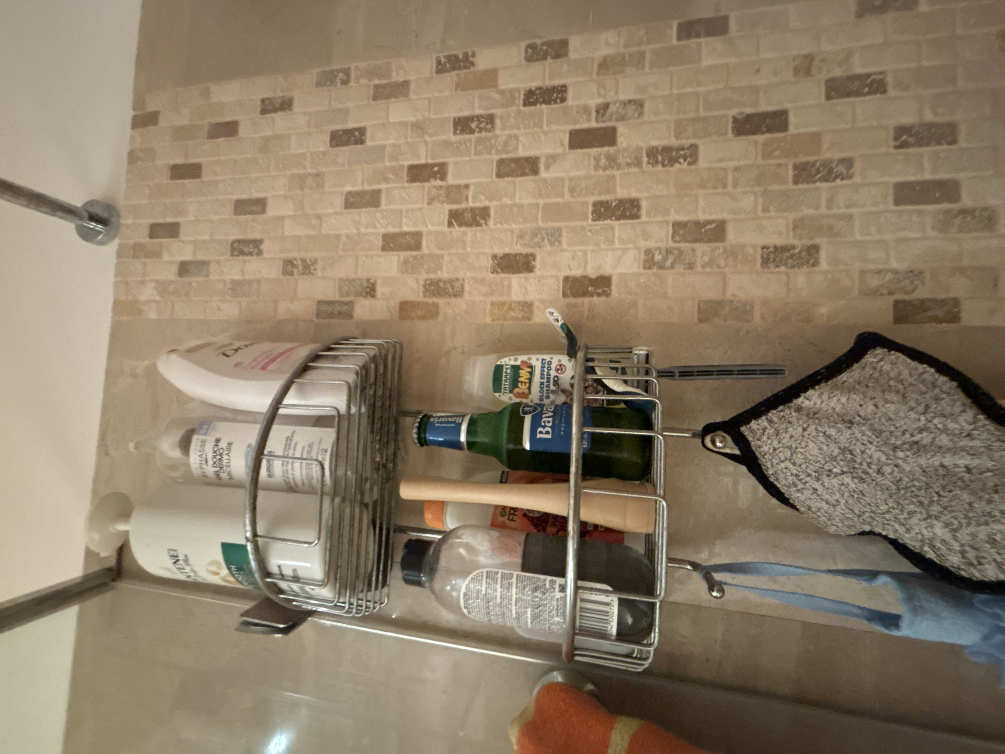 In a tiled shower stall, a metal shower caddy hangs from a showerhead pipe. The caddy contains various toiletries, including shampoo bottles, a toothbrush, and a green beer bottle. On the lower level of the caddy, a large clear bottle with a black cap is visible, along with a wooden object and a beer bottle. A blue bath pouf hangs from a hook below the caddy, next to a gray washcloth. The shower stall is adjacent to a tiled wall with a mosaic pattern of rectangular tiles in varying shades of beige and brown.