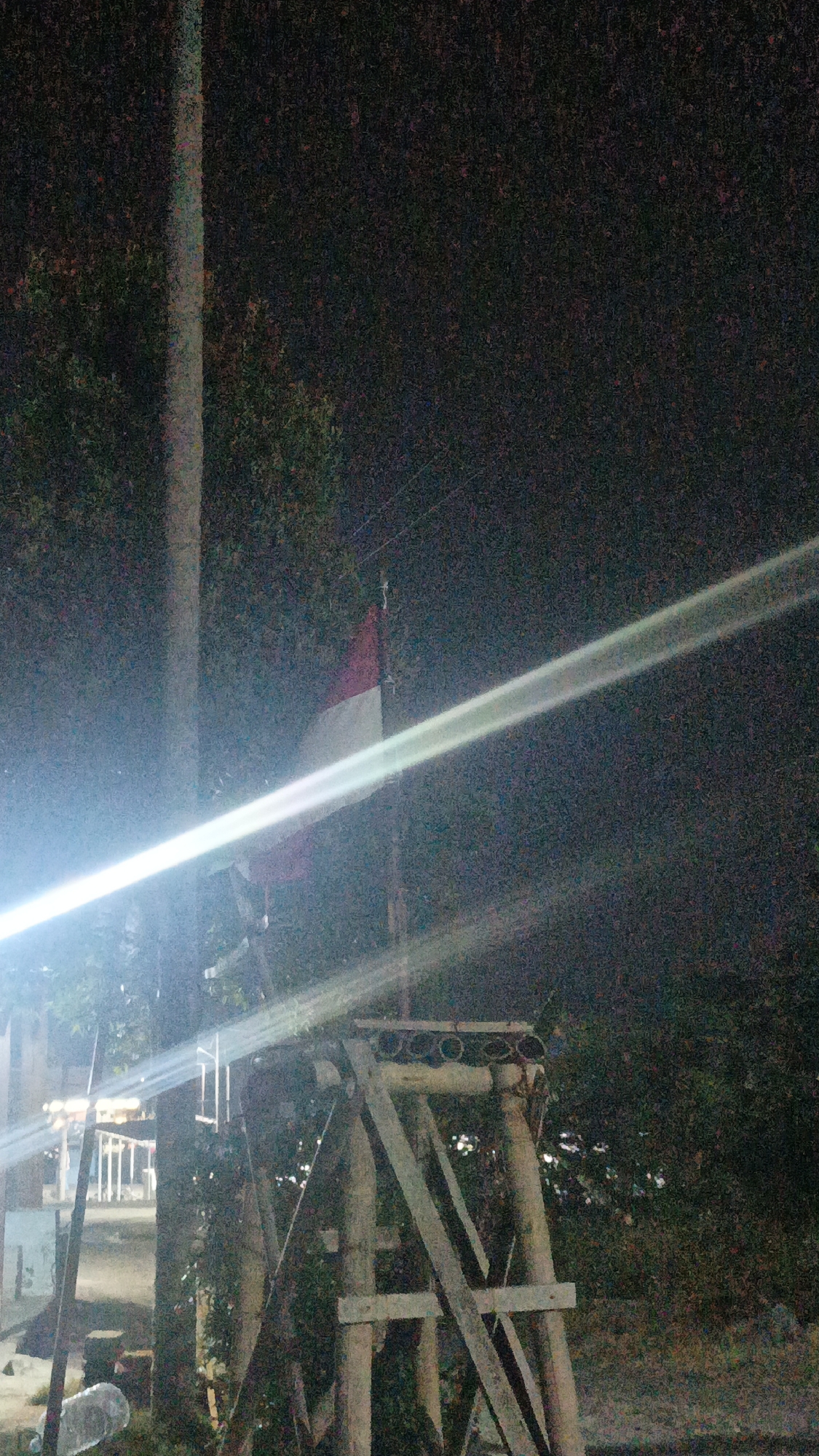 The image is a nighttime, low-light photograph taken outdoors, likely in the city of Sambi, Indonesia. A prominent feature is a tall, slender pole, possibly a utility pole, on the left side of the frame, illuminated by a bright, horizontal light streak that appears to be lens flare. Behind the pole, an Indonesian flag, displaying its red and white horizontal bands, is visible, attached to a flagpole. To the right of the pole and flag, a wooden structure, appearing to be a makeshift stand or support, is visible. The background consists of dark foliage and trees, with some faint lights suggesting distant buildings or streetlights. The ground is covered with debris, including a plastic bottle. The overall atmosphere is dark and somewhat grainy due to the low light conditions.