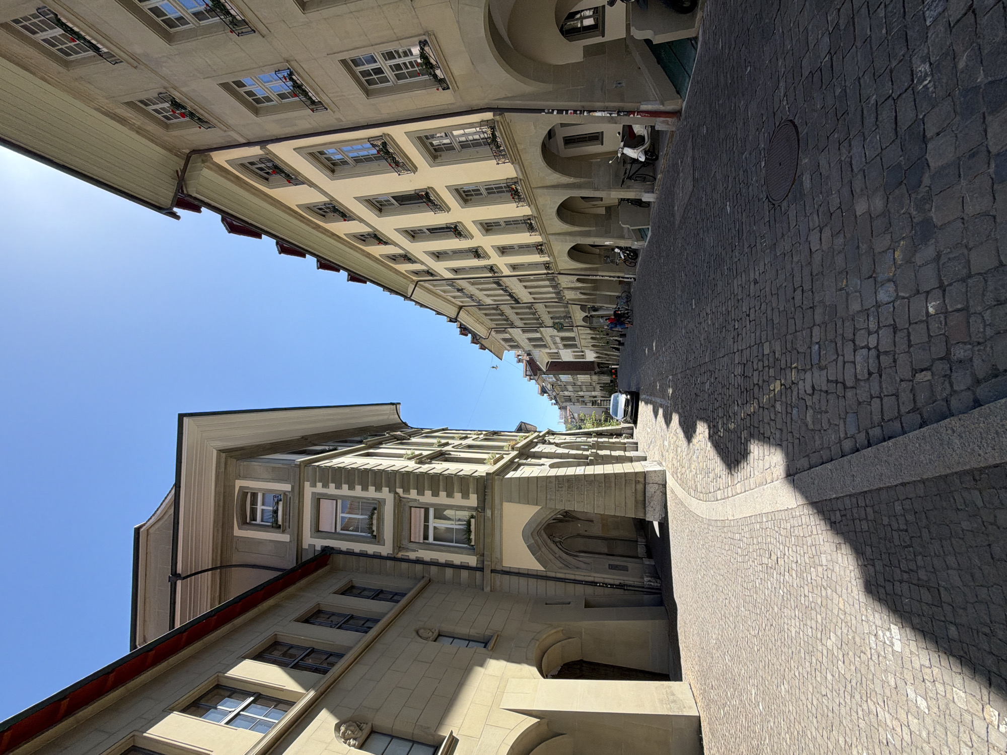 The image shows a narrow, cobblestone street in Bern, Switzerland, lined with historic buildings. The architecture features arcades on the ground floor and multiple stories with uniformly spaced windows. The buildings are constructed of stone, with light-colored facades and dark-tiled roofs. The sky is clear blue.
On the left side of the street, a building with arched entrances and stone ornamentation is prominently featured. The cobblestones in the foreground cast long shadows from the sun, indicating a sunny day.
On the right side, a continuous row of buildings with arcades creates a sheltered walkway. Several parked bicycles and scooters are visible, along with a white scooter in the foreground. A silver car is parked further down the street on the right. A few individuals are walking and interacting with their bicycles in the distance on the right side of the street.
The perspective of the image is a low-angle, wide shot, looking down the street, which curves gently to the right and disappears into the distance. The sun is high in the sky, creating bright lighting conditions. The overall scene depicts a charming, historic urban environment.