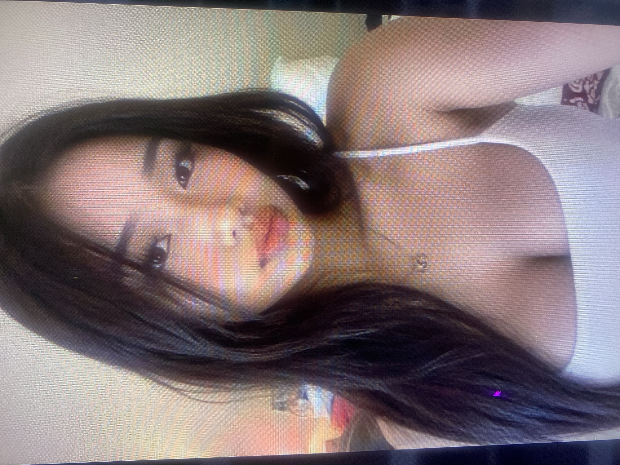 A close-up, pixelated image shows the upper body of a young East Asian woman. She has dark, long hair framing her face, which is lightly made up. She is wearing a white tank top and a delicate necklace with a pendant. The background is blurred and appears to be an indoor setting, possibly a bedroom, with hints of a pillow and some objects on a surface. There is a faint purple-pinkish light reflection on the left side of her hair. The image is too low resolution and lacks any discernible details to determine its location in Samara, Russia or any specific context.