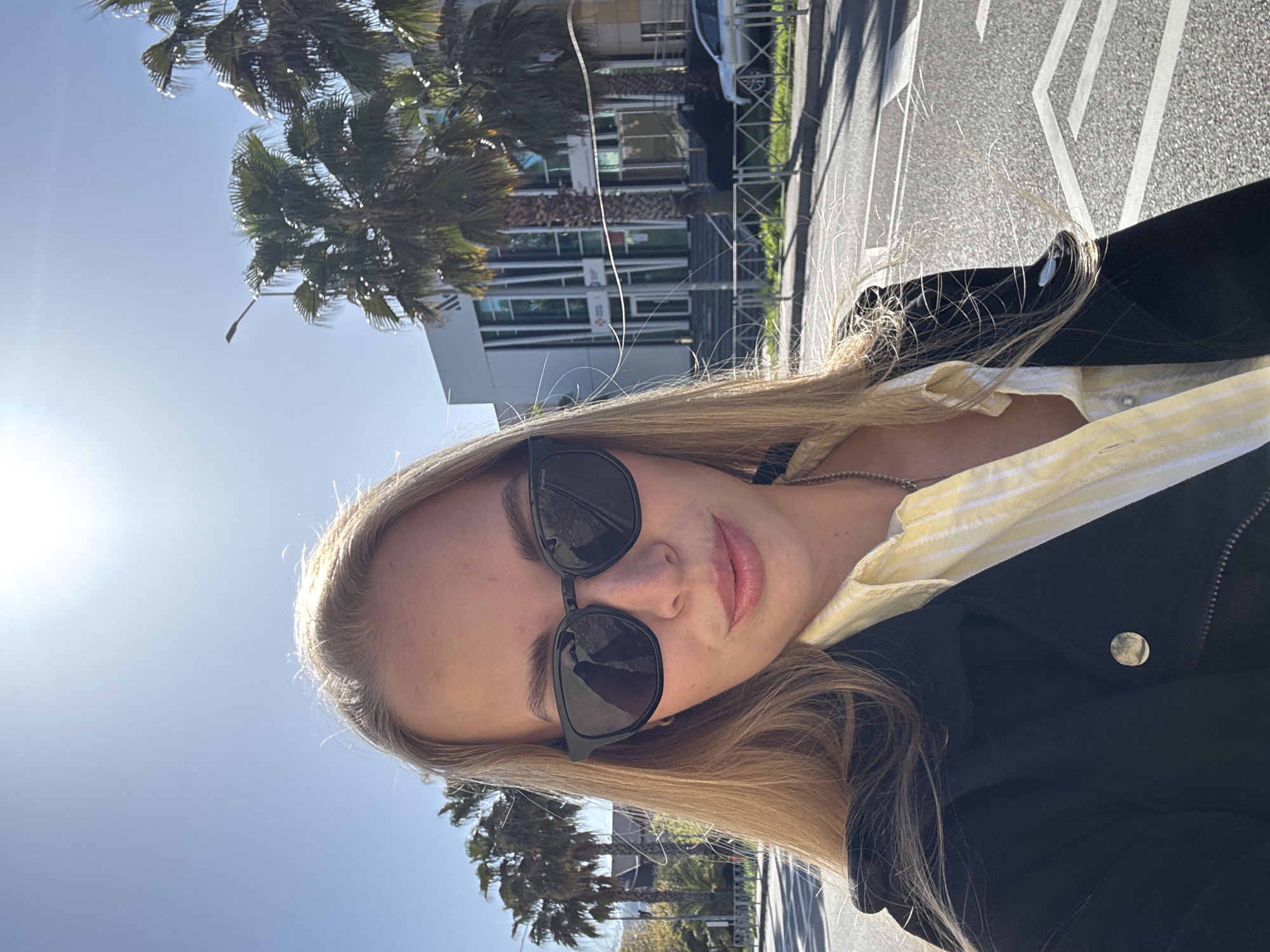 This media file presents a sunlit outdoor selfie of a young woman taken in Sochi, Russia. The image is oriented vertically, showing the woman's head and upper torso from a low angle, looking slightly upwards and to her left. Her long, blonde hair frames her face, and she wears black sunglasses, on which the brand "Polaroid" is visible on the side. Her expression is neutral, with slightly parted lips. She is dressed in a black jacket featuring a visible zipper and a round, metallic button-like detail, over a yellow and white striped shirt. A silver-colored necklace is also visible.
The background depicts a bright, clear blue sky with the sun shining intensely from the upper left, creating a prominent lens flare. Several palm trees with dark green fronds are scattered across the mid-ground, some catching the direct sunlight. Behind the foliage, a modern building with multiple windows and light-colored structural elements can be seen, and a light pole stands against the sky. To the right side of the frame, a paved road with clear white markings, possibly directional arrows, extends into the distance. A silver car is partially visible near the building. The overall scene conveys a bright and clear day in an urban or developed setting.