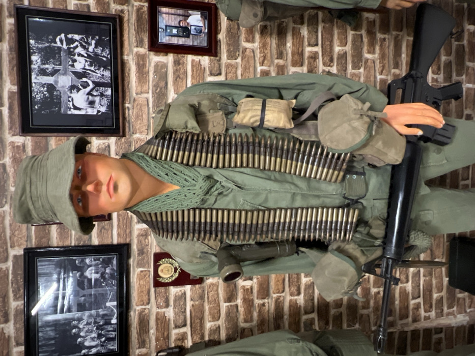 A mannequin dressed in a green military uniform stands against a brick wall. The mannequin is wearing a bucket hat and has ammunition belts draped across its chest. It holds a black rifle. Behind the mannequin are three framed pictures and a small medal.