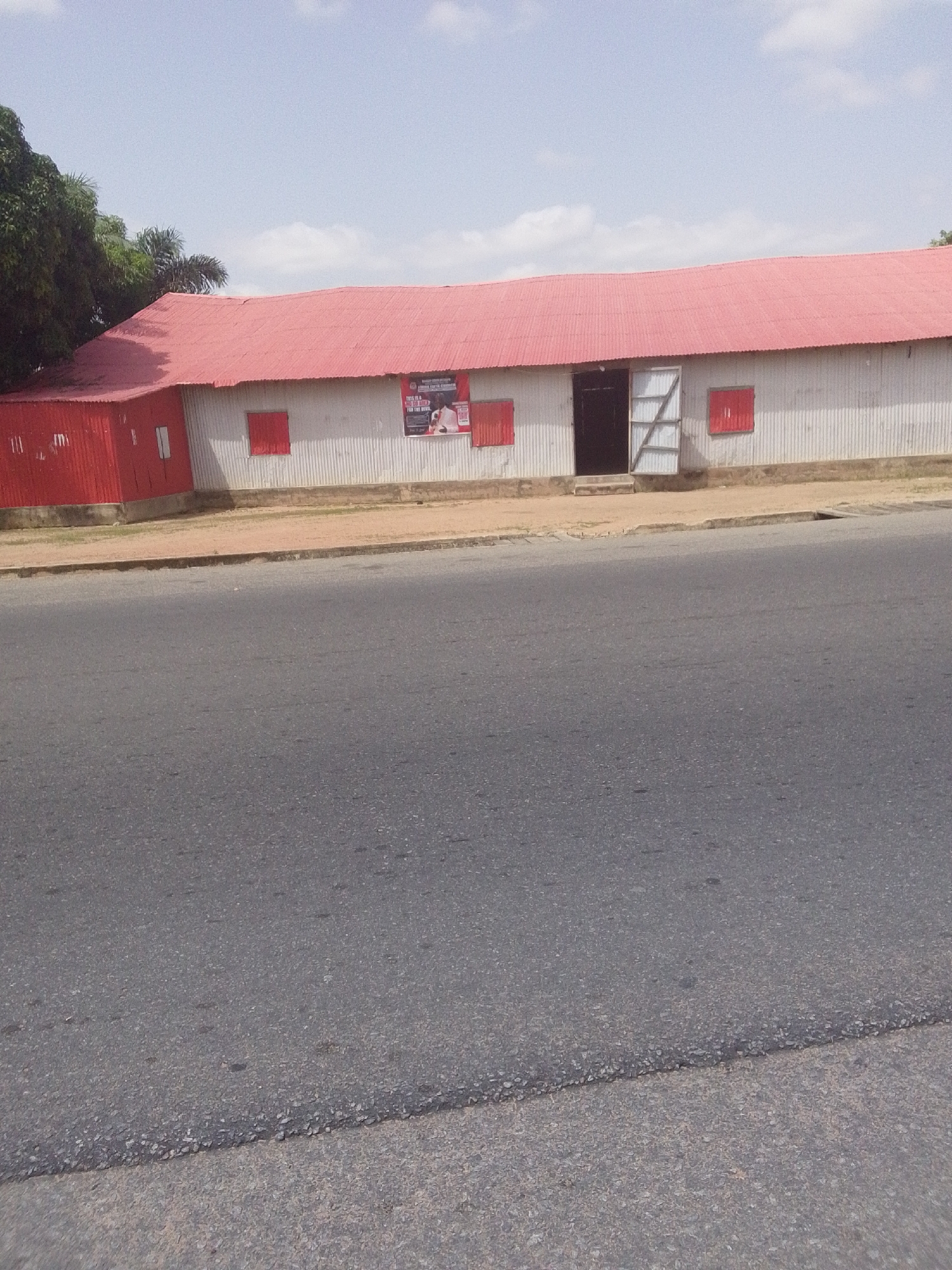 A long, low-lying building with a red corrugated metal roof and corrugated metal siding is situated beside a paved road. The building has several red-shuttered windows and a large doorway with double doors, one of which is open. A poster featuring a man in a suit is affixed to the wall near the doorway. To the left of the main building, a section of red corrugated metal wall extends outwards. Green trees are visible behind the building on the left. The scene appears to be in an urban or semi-urban environment in Jalingo, Nigeria, based on the building style, vegetation, and road condition. The sky is partially cloudy.