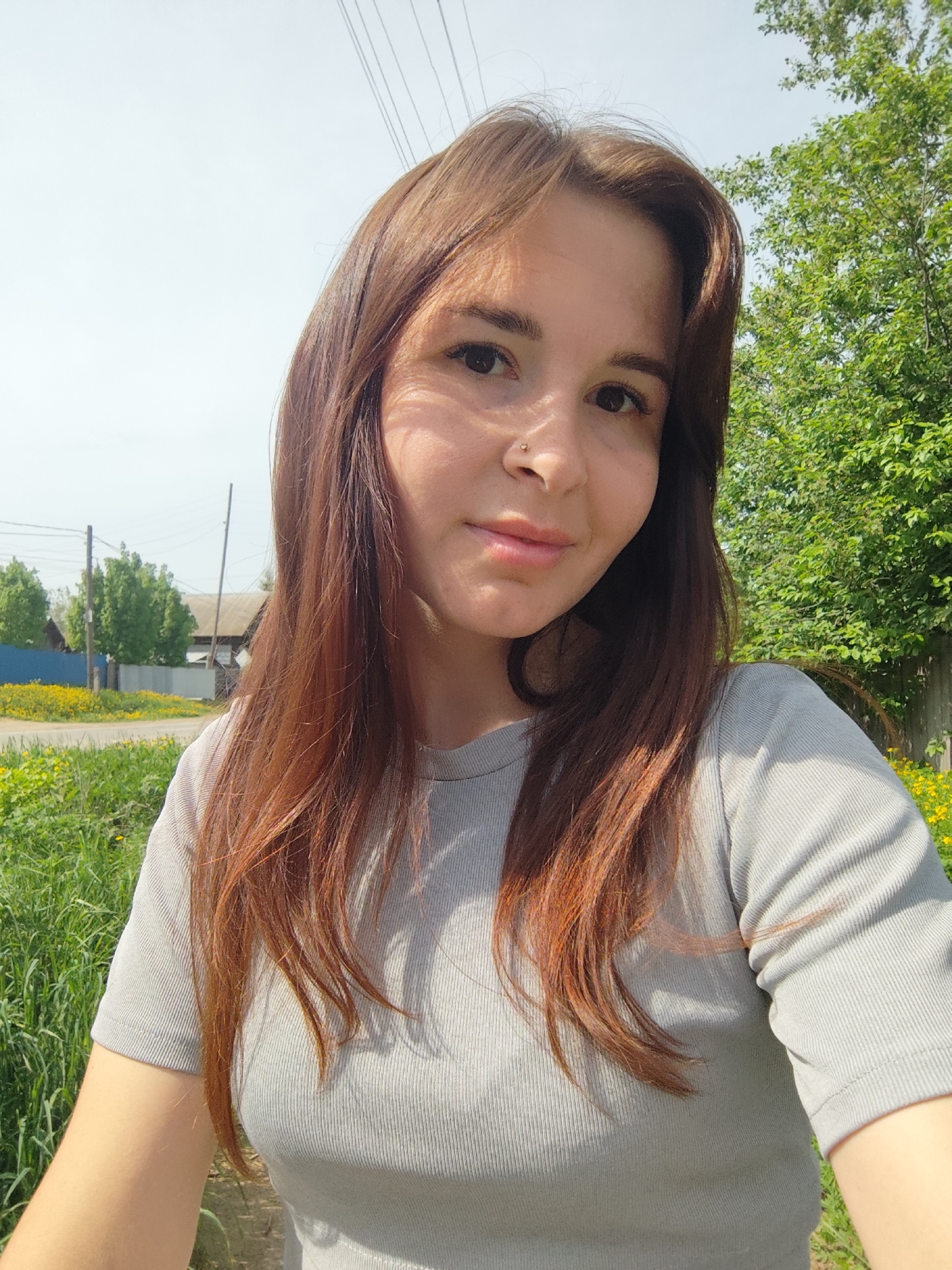 A young woman with long, auburn hair poses for a selfie outdoors. She wears a gray t-shirt and has a nose piercing. The background includes a grassy area with yellow wildflowers, a road, a wooden fence, and residential buildings. Telephone poles and trees are also visible in the scene. The photo appears to have been taken in daylight with natural lighting. The location is near Фоки, Russia.