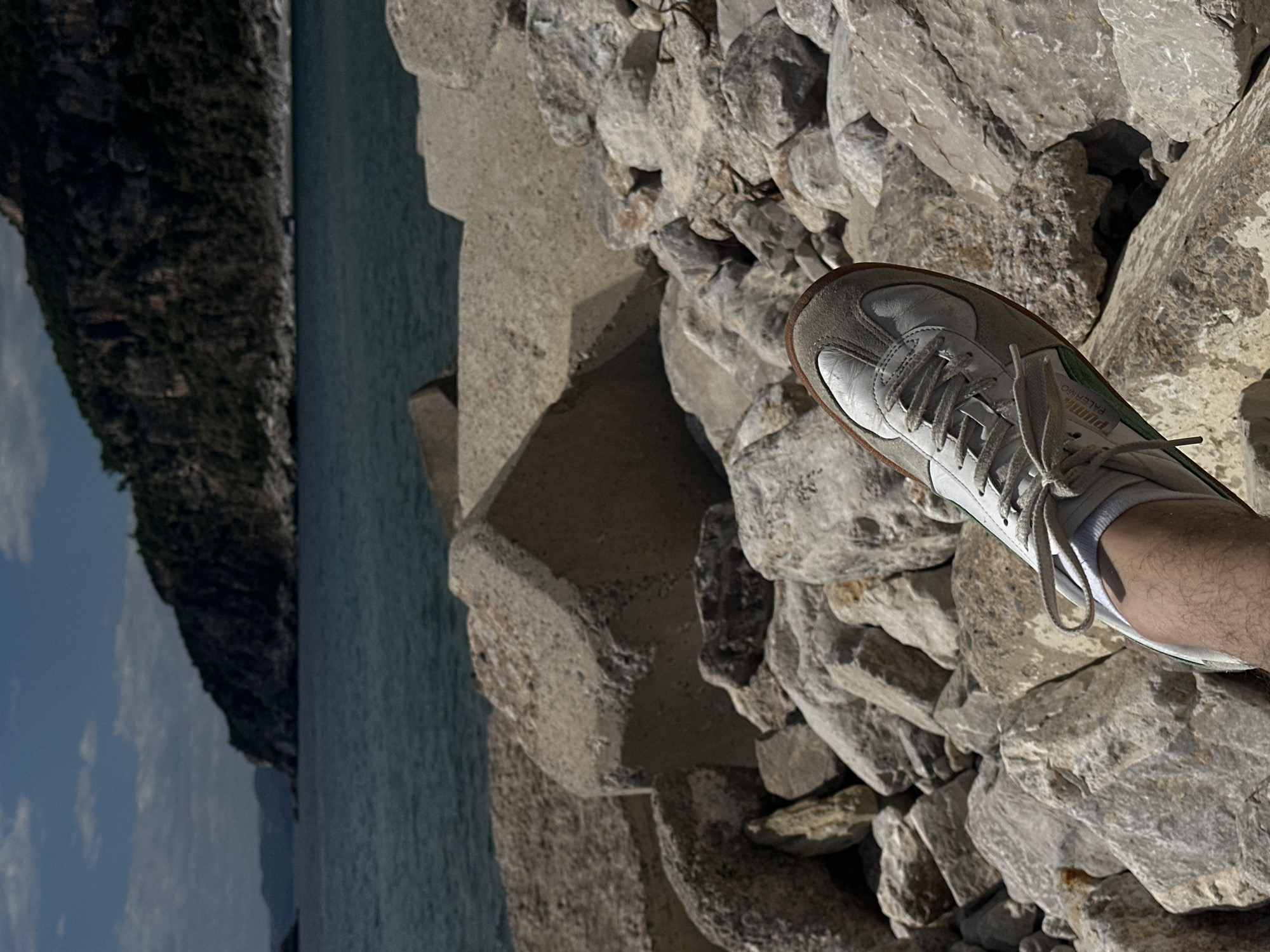 A person stands on a rocky cliff face in Perazića Do, Montenegro. The person's right foot, clad in a white sneaker, is placed on the rocks. Beyond the cliff face is the Adriatic Sea, with tree-covered mountains in the background. The sky is partly cloudy.