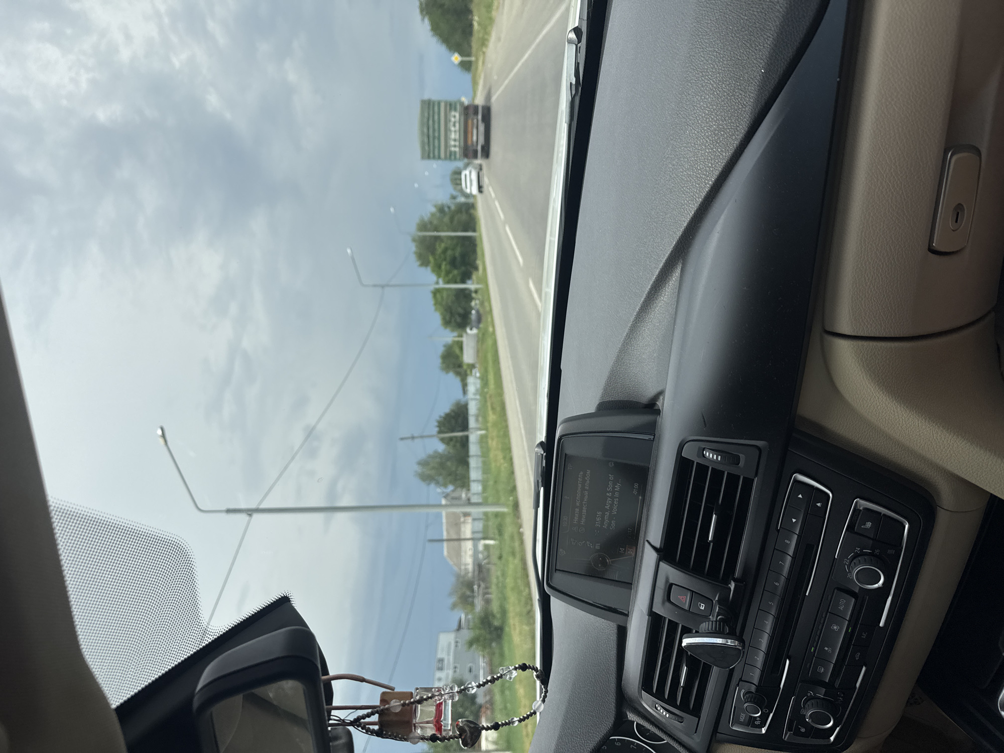 The image is a perspective shot from inside a car, looking out at a highway with a truck and a white car ahead. The car's dashboard is visible in the foreground, with its entertainment screen displaying music information. The sky is overcast and grey, suggesting an impending storm or a generally cloudy day. The road is paved and appears to be in good condition, with clear lane markings. On either side of the road, there is grass, trees, and some distant buildings. The overall scene depicts a journey on a highway, likely in a rural or semi-rural setting. Based on the context provided, the location is the city of Novotroitskaya, Russia.