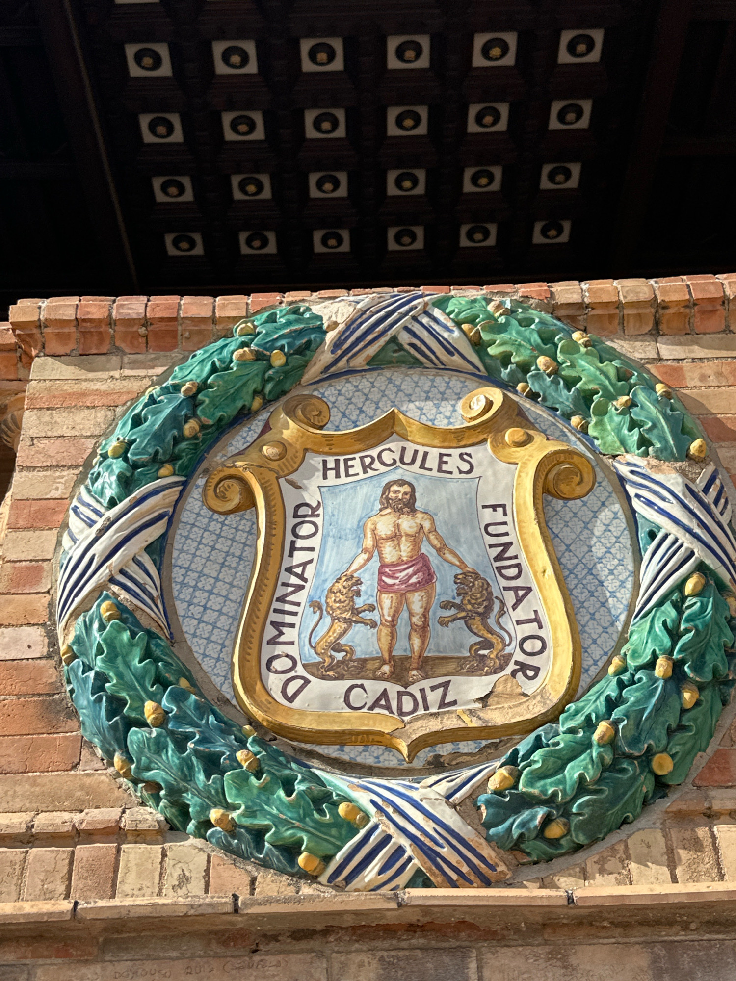 The image shows a detailed ceramic crest mounted on a brick wall. The crest is circular and framed by a wreath of green oak leaves and acorns, with interlaced blue and white ribbons. At the center of the crest is a shield bearing a depiction of Hercules, a muscular, bearded male figure with a red cloth around his waist, standing between two lions. Above Hercules, the word "HERCULES" is inscribed, and on either side of him, the words "DOMINATOR" and "FUNDATOR" are written in a semi-circle. Below Hercules, the word "CADIZ" is inscribed on a banner. The crest is located on the exterior wall of the Plaza de España in Seville, Spain. The background shows a dark, vaulted ceiling with a grid of square elements, each containing a circular golden disc.