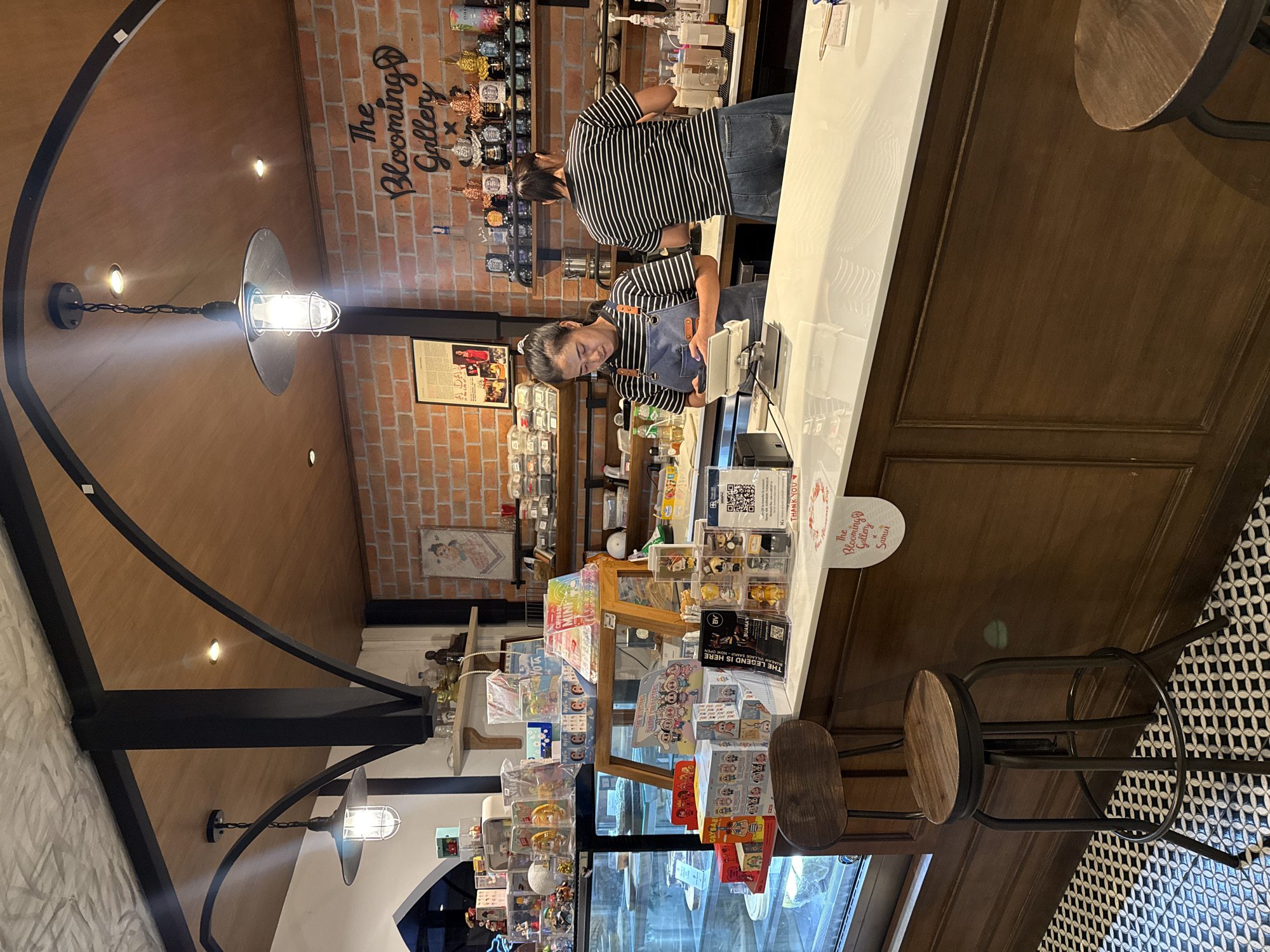 This image depicts the interior of a cafe or gallery located in Ko Samui District, Thailand. The scene is viewed from the front, looking towards a counter area. Two female employees are visible behind the counter. One, wearing an apron and a black and white striped shirt, is interacting with a tablet device. The other employee, in a black and white striped shirt and blue jeans, is facing away from the camera and appears to be preparing drinks.
The cafe's interior features exposed brick walls, wooden ceiling beams, and industrial-style pendant lights. Shelves behind the counter are stocked with various items, including decorative figures, packaged goods, and what appear to be small bottles or containers. A glass display case runs along the left side of the counter, showcasing more items, possibly food or merchandise.
In the foreground, a rustic wooden stool with a metal frame is positioned next to the counter. The floor is covered with a patterned black and white tile. A circular sign with text that includes "The Blooming Gallery" and "Samui" is placed on the counter. Several bottles and dispensers are visible on the counter, suggesting a beverage service area. The overall impression is a well-lit, stylishly decorated establishment.