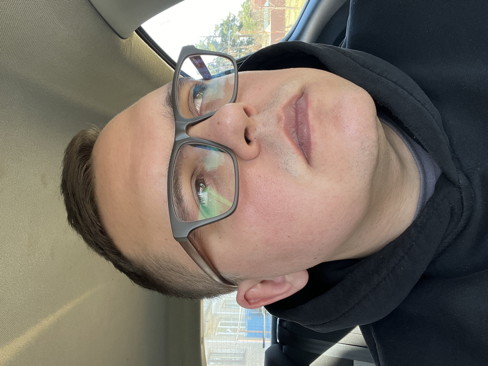 A close-up, portrait-style image captures a young adult male with short dark hair, light skin, and green eyes, wearing dark-rimmed rectangular glasses. His head is tilted significantly to the left (counter-clockwise) within the frame. He has a neutral facial expression with his lips closed. Some light stubble is visible on his chin and upper lip area.
The man is wearing a black hooded sweatshirt over a grey undershirt. He appears to be inside a vehicle, with parts of a light-colored, textured car headliner visible in the upper left of the frame. Through the car windows, external scenery is visible, including hints of green trees and portions of a building featuring light-colored walls and blue accents.
The scene takes place during the daytime, suggested by the bright outdoor light and clear reflections in his glasses. The reflections show elements of the sky and foliage. No visible text is present in the image. The implied location context for this media file is Раздолье, Russia.