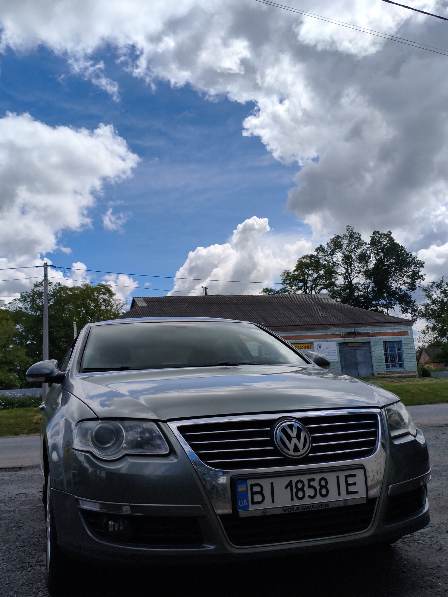 A silver Volkswagen sedan is parked on the side of a road. The license plate reads "BI 1858 IE" and has a Ukrainian flag on it. Behind the car is a one-story building with a gray roof and blue and white facade. Trees are visible behind the building. The sky is blue with fluffy white clouds. The car is likely parked near a building in Ustyvytsia, Ukraine.