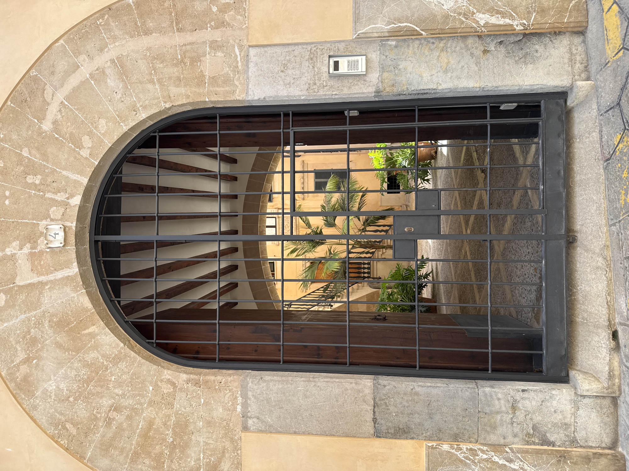 The image shows a view through an open arched doorway, which is secured by a dark metal gate. The doorway is framed by a rough-hewn stone arch and stone pilasters. Above the arch, a white square plaque with the number "4" is affixed to the building's facade, which is painted a pale yellow.
The gate itself is made of vertical and horizontal bars, forming a grid pattern. Through the gate, a sunlit courtyard is visible. The courtyard features a palm tree in the center, surrounded by potted plants. A staircase with a decorative metal railing leads up along the left side of the courtyard. The buildings surrounding the courtyard are painted in a warm, yellow hue, and some windows with dark shutters are visible. A light-colored intercom system is mounted on the stone pilaster to the right of the entrance. The ground in front of the doorway is paved with hexagonal stones, with yellow lines painted on some of them.
This scene is located in Palma de Mallorca, Spain. Specifically, this is the entrance to a courtyard located at Carrer de Sant Miquel, 4, in Palma de Mallorca.
