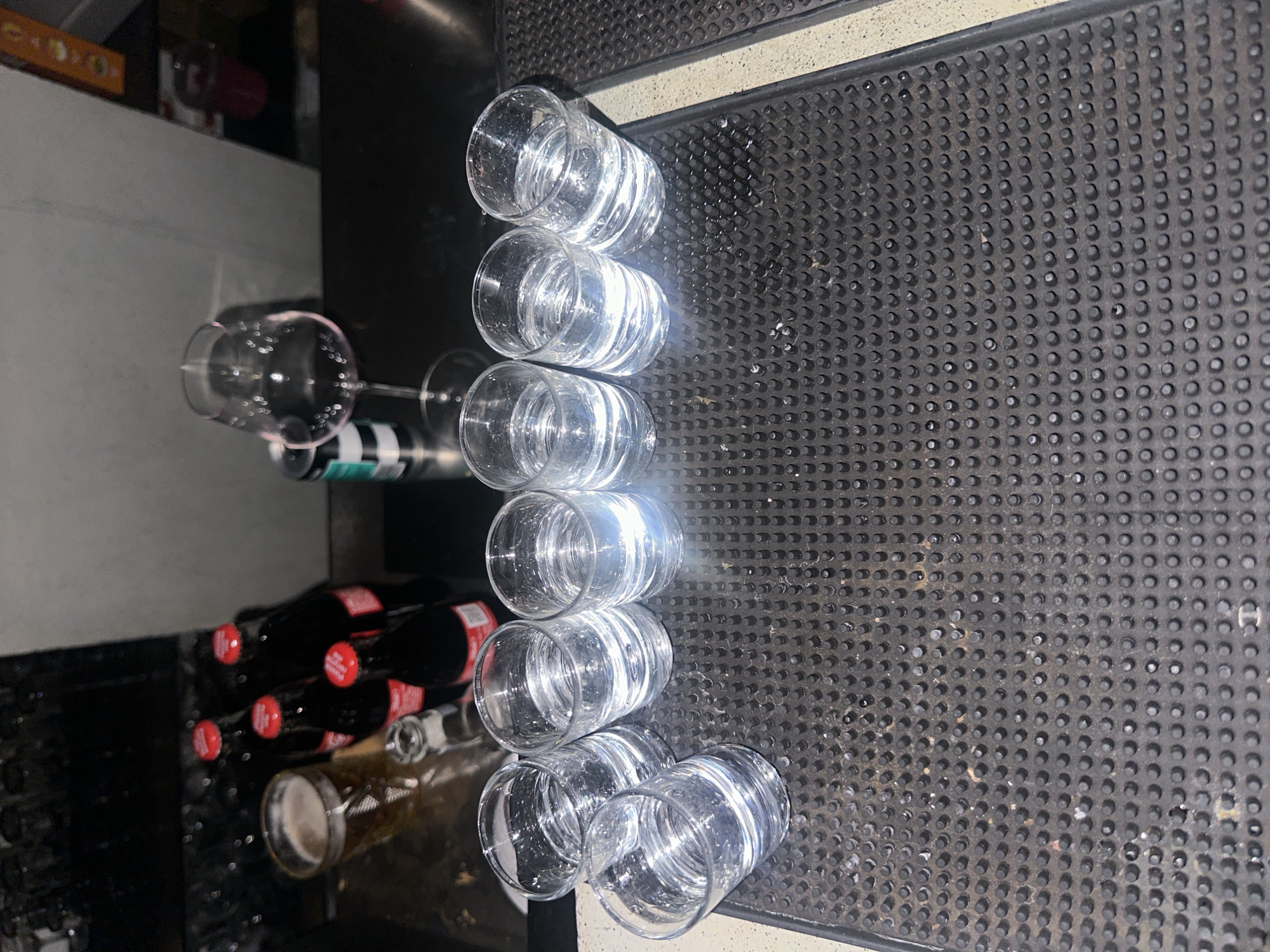 The image shows a bar setting. In the foreground, a textured black rubber mat is visible with circular indentations throughout the surface. Arranged on this mat are a row of clear, empty glasses stacked one behind the other. Eight of the glasses appear to be tumblers or similar types for serving drinks. Behind the glasses, to the left, there are partially visible bottles of what appear to be alcoholic beverages, some with red bottle caps. A wine glass is also present, lying on its side. The location is a bar, but the details do not indicate that it is in London, United Kingdom.