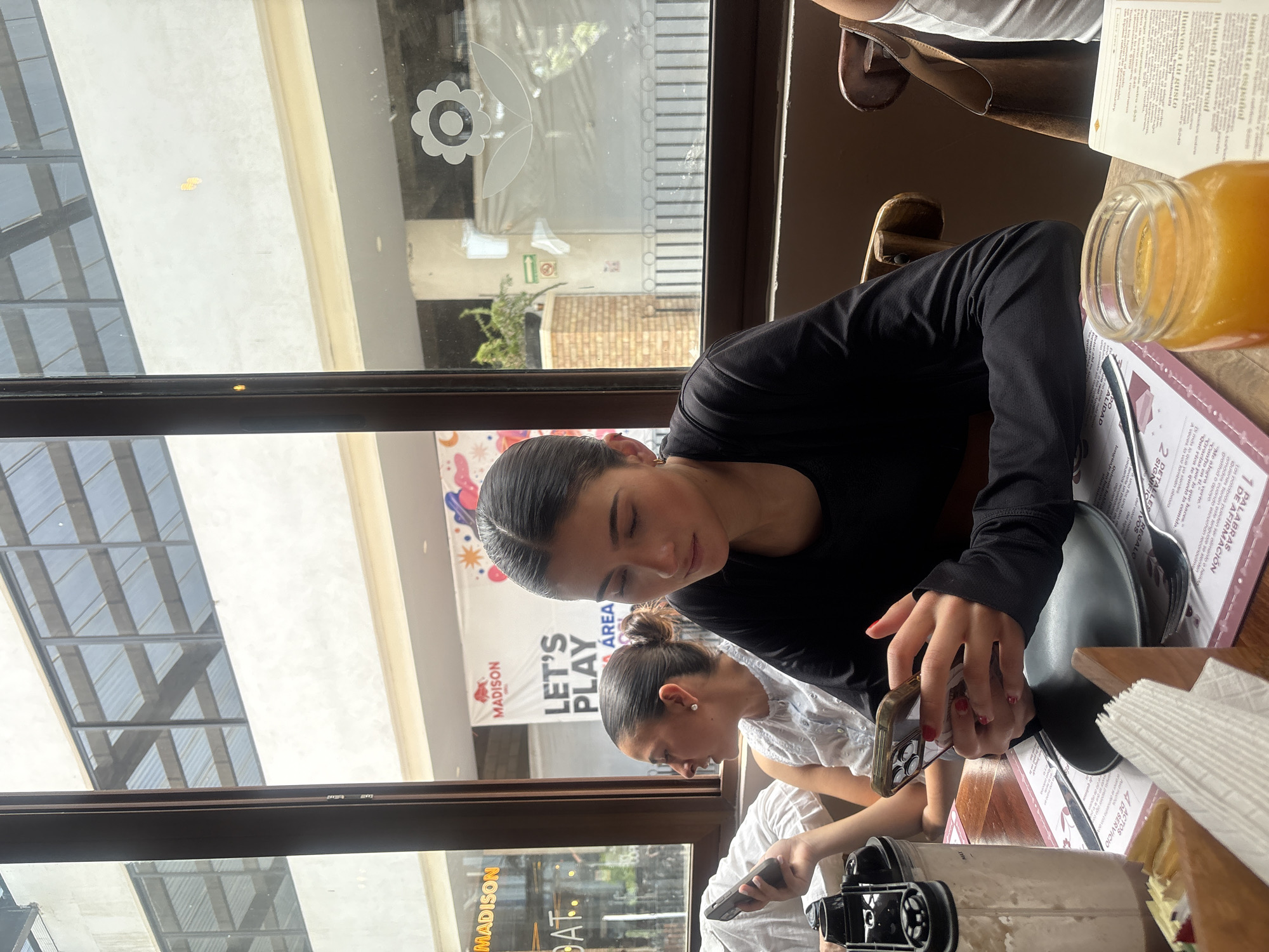 The image captures an indoor scene at a restaurant or café, possibly in Puebla, Mexico, as "MADISON" is prominently displayed outside. Two women are seated at a table near a window. One woman, wearing a black long-sleeved top, is handling a mobile phone with red nail polish, while the other woman, wearing a white top, looks on. The table contains menus, a plate, a glass jar filled with yellow liquid (likely juice), utensils, and an object that appears to be a water filter. Through the window, a view of an exterior building and the street is visible.