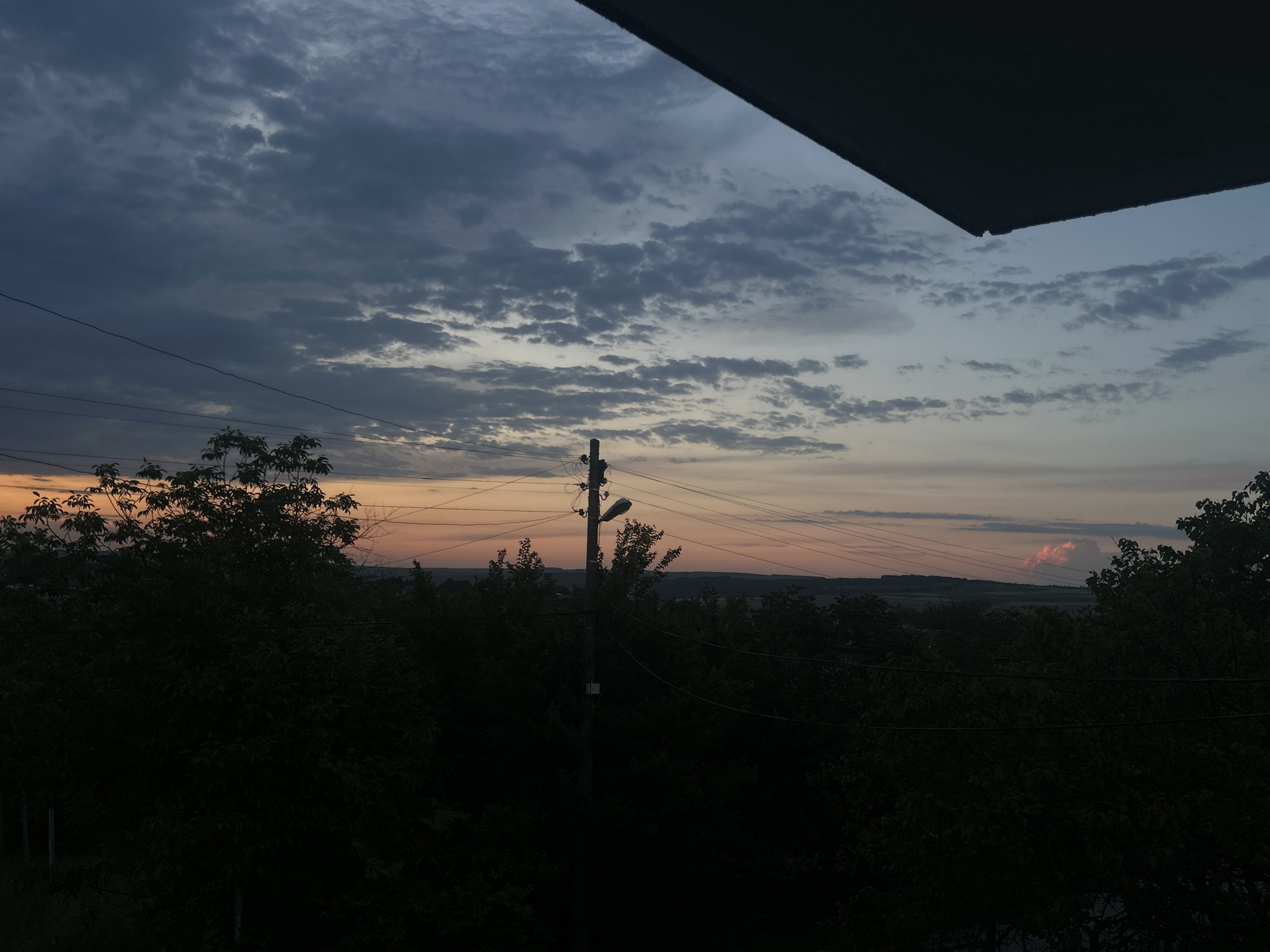 The image shows a view of a sunset from what appears to be a high vantage point, possibly a balcony or window. The sky is filled with clouds in various shades of gray and blue, with hints of orange and pink near the horizon. A dark object, possibly part of a roof or structure, is visible at the top of the frame. Below the horizon line, there is a dense collection of trees and vegetation. A utility pole with wires is positioned in the middle ground, appearing to be part of the landscape. The scene is set in a location that allows a wide view of the landscape, with Loznitsa, Bulgaria being the specific location.