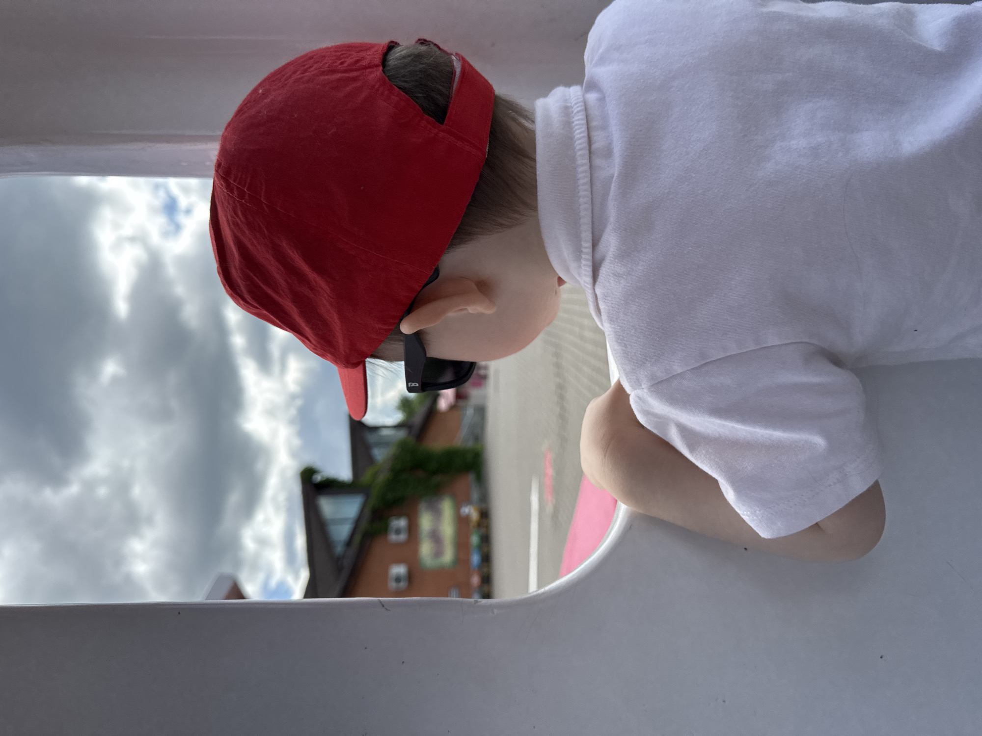 The image displays a young child, viewed from behind, situated within a white, curved structure, potentially part of a ride or play equipment. The child is wearing a red baseball cap, black sunglasses, and a white short-sleeved collared shirt. Their right arm is visible, bent at the elbow, resting on the white structure. The child is looking out through an opening in the structure towards an outdoor scene.
In the background, a paved area extends towards a reddish-brown brick building with a dark roof and prominent glass window sections. Green vines cling to a portion of the building's exterior. Several rectangular units, possibly air conditioners or signs, are mounted on the building's facade. A line of colorful, small vehicles, consistent with children's amusement rides, is visible on the paved surface in front of the building. The sky is predominantly overcast with gray clouds, though a small area of blue sky is discernible in the upper left. A subtle pink surface is visible immediately beyond the white structure in the foreground. No specific visual markers within the image provide confirmation of the location as Izhevsk, Russia.