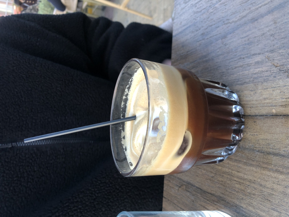 The image shows a glass of iced coffee on a wooden table. There is a black metal straw in the coffee. The coffee is made up of two layers: a darker coffee layer on the bottom and a lighter layer of milk foam on top. The glass has a fluted bottom and is sitting on a table with a wooden surface. The table is brown and has a slightly rough texture. The coffee is partially obscured by a black jacket, which is partially visible in the upper left corner of the image. The location is a cafe in Palaio Faliro, Greece, based on the given latitude and longitude. There is no person visible in the image. It is a still-life. The person's arm is visible in the background. The camera is at a slightly low angle and the subject is in focus. The image is well-lit.