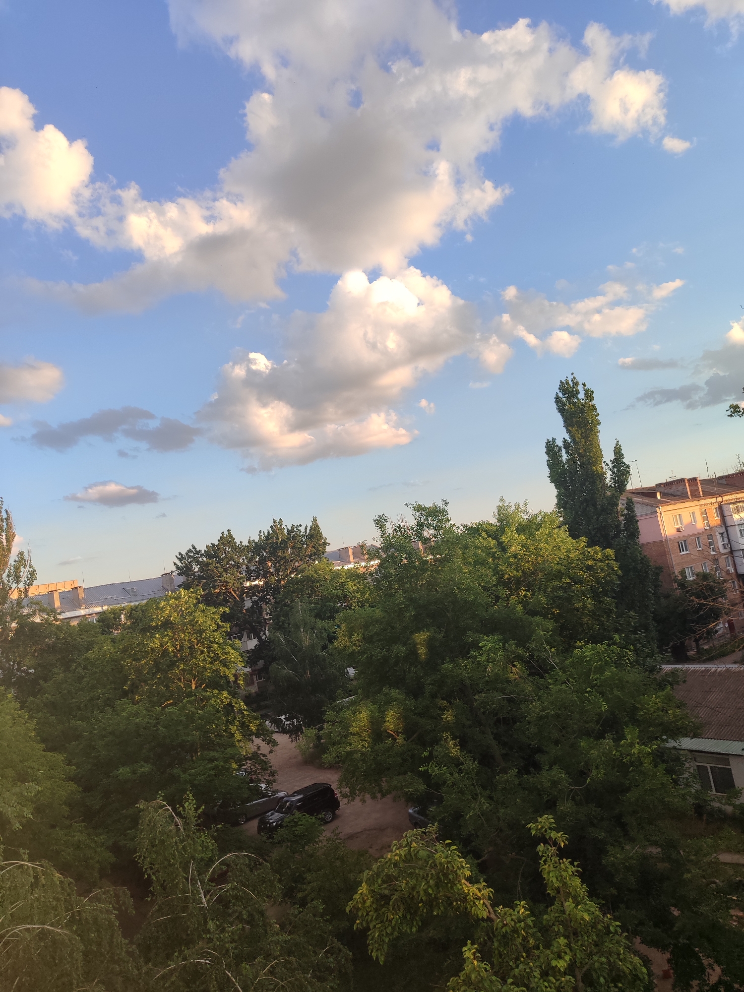 This is an outdoor, high-angle shot of a city scene in Mykolaiv, Ukraine. The sky is a clear blue with scattered white and light gray clouds, some of which are tinged with yellow from the sun. Below the sky are numerous trees with dense green foliage, some reaching considerable heights. Interspersed with the trees are several buildings.
In the foreground, on the left, is the top of a light-colored, leafy tree. Further into the scene, a cluster of various trees dominates the view, with some appearing taller and more slender than others. Behind the trees on the right side of the image, a multi-story residential building with a pinkish-orange facade and white-framed windows is visible. The building has a reddish-brown tiled roof. To the left of this building, another, longer building with a light-colored facade and a grey, sloped roof is partially obscured by trees.
In the middle ground, between the trees and the buildings, a parking area is visible. Several dark-colored cars are parked on a dirt or gravel surface. One black SUV and a dark sedan are clearly discernible.
The overall impression is that of a relatively urban environment with significant green spaces, likely a residential area with mature trees providing substantial canopy cover. The lighting suggests it is late afternoon or early evening, given the warm tones on some of the clouds and the dappled sunlight on the foliage. There are no people visible in the image.