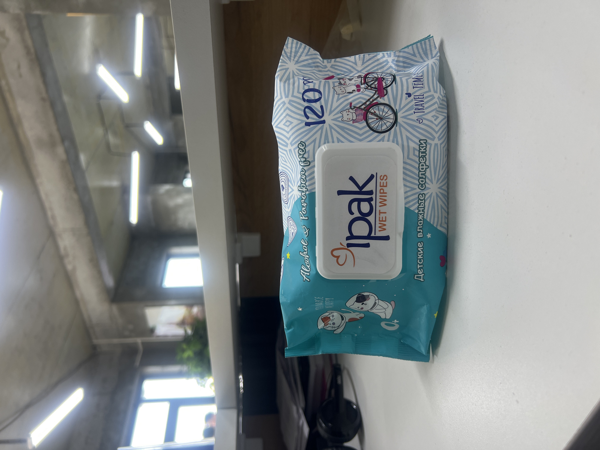 A close-up, eye-level, medium shot features a package of "ipak WET WIPES" positioned on a white, flat surface, possibly a desk. The packaging is predominantly teal and white, adorned with a repeating diamond pattern in light blue. Graphics on the package include two cartoon cats, one wearing a bow, and another pair of cats riding a bicycle. Text on the packaging indicates "120 PCS," "Alcohol & Paraben free," and "TRAVEL TIME." Below the main branding, the phrase "Детские влажные салфетки" is visible in Russian, translating to "Baby wet wipes." The background is blurred, revealing an indoor environment with concrete walls and ceiling, fluorescent lighting fixtures, and windows with natural light. A hint of greenery is visible through one of the windows, suggesting plants. The overall scene is indoor and office-like. There are no people or animals present in the immediate vicinity of the product. The image does not contain any identifiable landmarks or features that would place it specifically in Shahrixon, Uzbekistan.