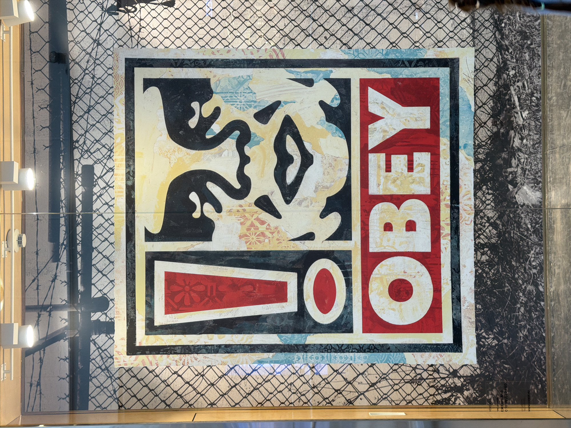 An Obey Giant street art poster is displayed on a glass storefront in Moscow, Russia. The poster features a stylized portrait of a face with the word "OBEY" in large red letters below it. The poster is behind a chain-link fence, and the background shows what appears to be a retail store interior with lighting fixtures. To the left of the poster, there is a sign in Russian that reads "KXBOX ЕЖЕДНЕВНО С 10:00 ДО 23:00" which translates to "KXBOX DAILY FROM 10:00 TO 23:00".
