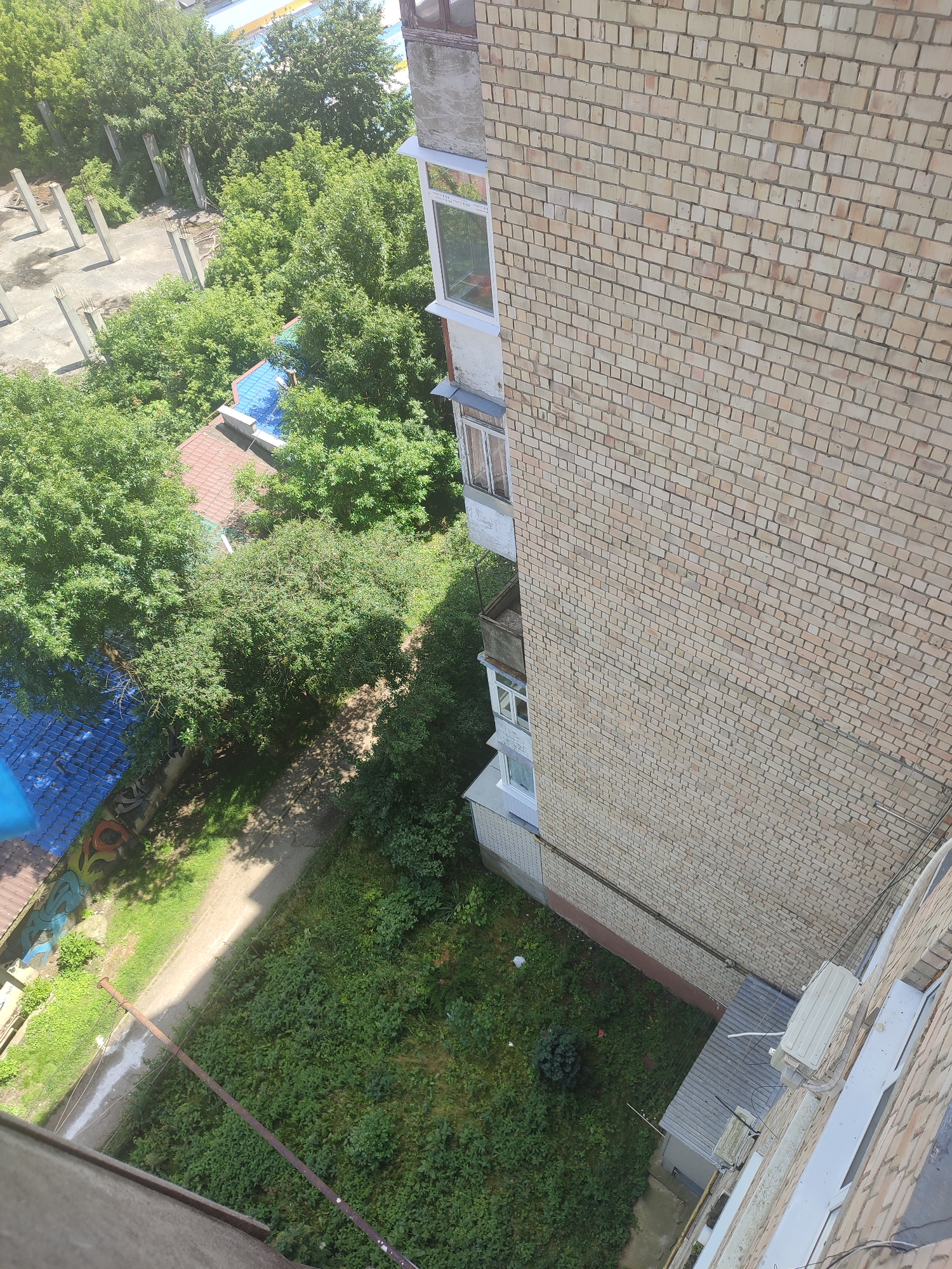The image is a high-angle, outdoor, daylight shot looking down on a courtyard area adjacent to a brick apartment building. The building's light-colored brick facade, featuring multiple windows and balconies, dominates the right side of the frame. In the foreground, the edge of a concrete structure, possibly a balcony or windowsill, is visible. Below the building, a densely vegetated area with various green plants and some scattered grass is present. A paved path runs through the vegetation, with a rusty metal pole running diagonally across it. To the left of the path, there is a section of ground with more grass and a small bush. Further to the left, a small building with a blue, corrugated roof is visible, along with a section of another building with a reddish-brown roof. There are also signs of construction, with several concrete pillars standing in a cleared area behind some trees. The overall impression is one of an urban environment with abundant greenery and some signs of development or recent construction. Based on the provided context, this scene is located in Chernivtsi, Ukraine.