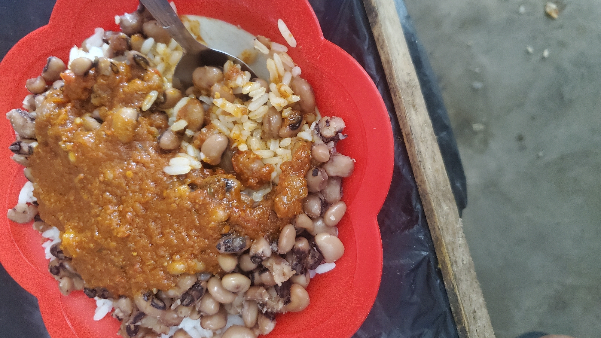 A close-up overhead shot shows a bright red, scalloped-edge bowl filled with a meal. The bowl contains white rice covered by a thick, reddish-brown stew and a generous portion of black-eyed peas. The stew has a coarse texture, and some of the peas are visible underneath it, with the majority surrounding the rice and stew. A silver spoon is partially submerged in the food on the right side of the bowl. The bowl rests on a black surface, possibly plastic sheeting, which is laid over a wooden plank. The background consists of a grey, textured surface, likely concrete. The image is lit from above, casting subtle shadows. The scene is likely from a food stall or eatery in Jalingo, Nigeria, given the presence of local cuisine and common food service materials.