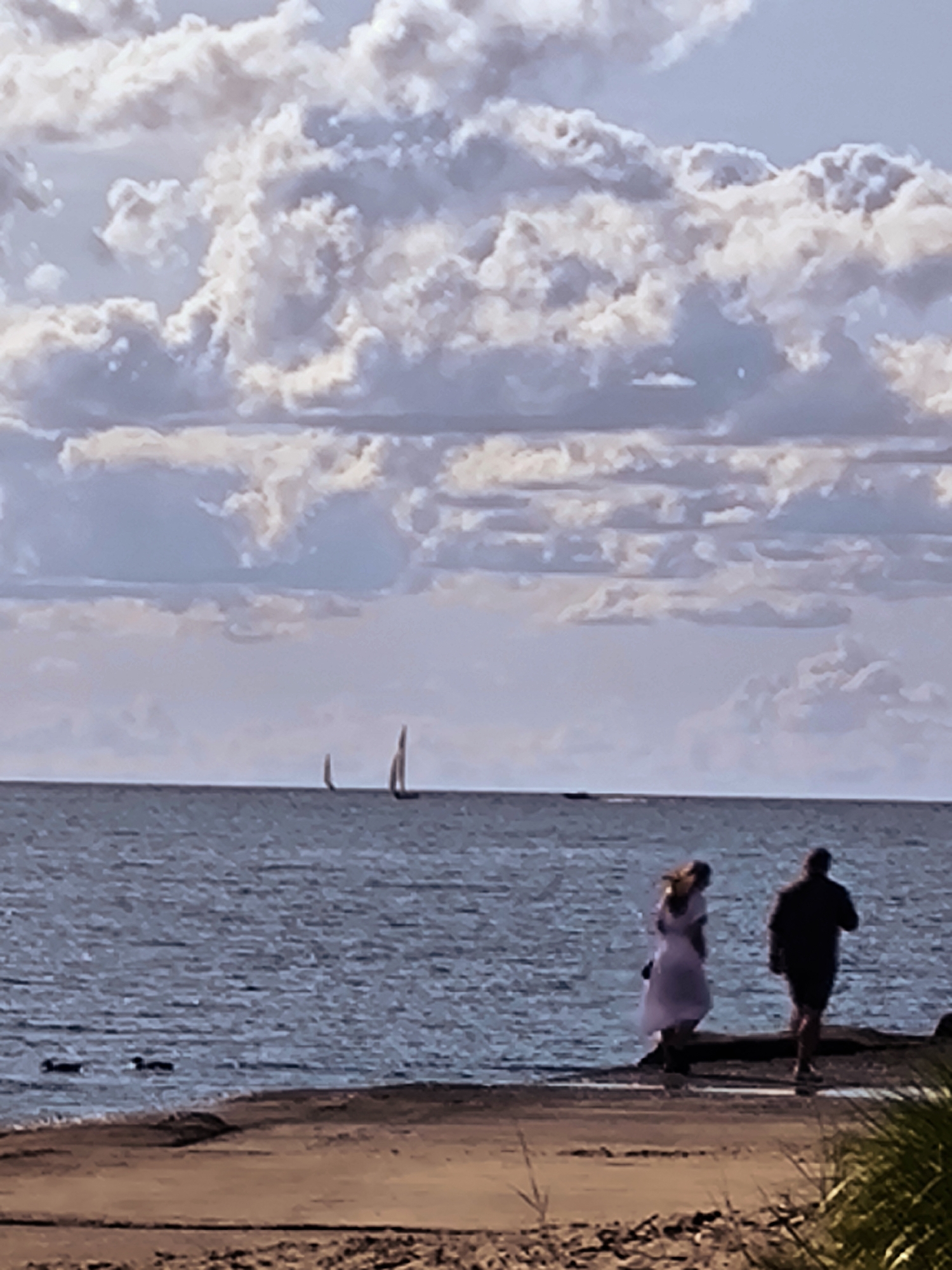 An outdoor, low-angle, medium shot captures a hazy view of a body of water with two sailboats on the horizon. In the foreground, two figures walk along a sandy shore. The person on the left is wearing a light-colored, flowing dress and has blonde hair. The person on the right is wearing a dark-colored shirt and shorts. The sky above is filled with large, dramatic clouds in shades of white and gray, with patches of blue sky visible. The overall impression is of a calm, overcast day at the waterfront, possibly near a park or beach area in Riga, Latvia.